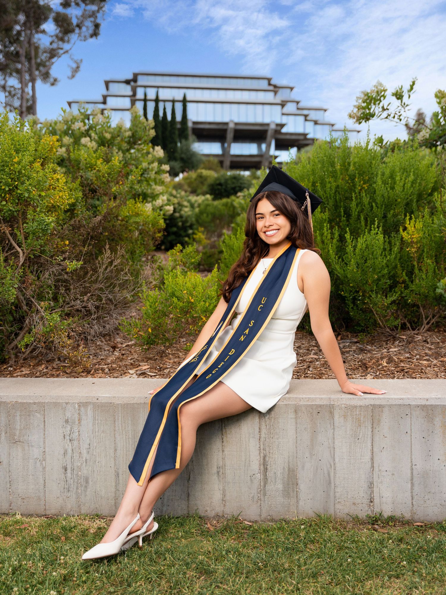 Graduate in white dress and academic stole sits on concrete ledge with architectural structure in background.
