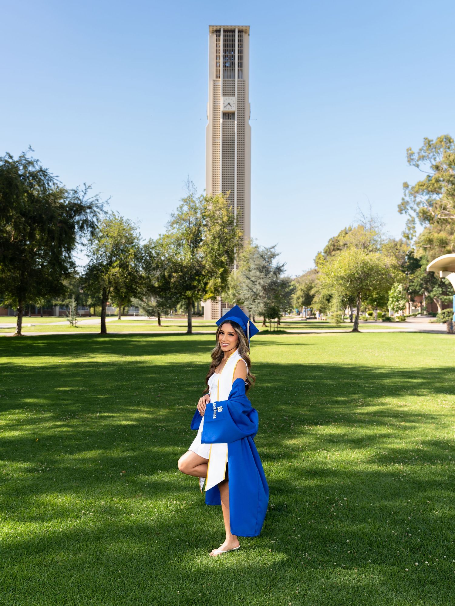 Woman in blue graduation cap and gown posing on green campus lawn with tall bell tower in background.