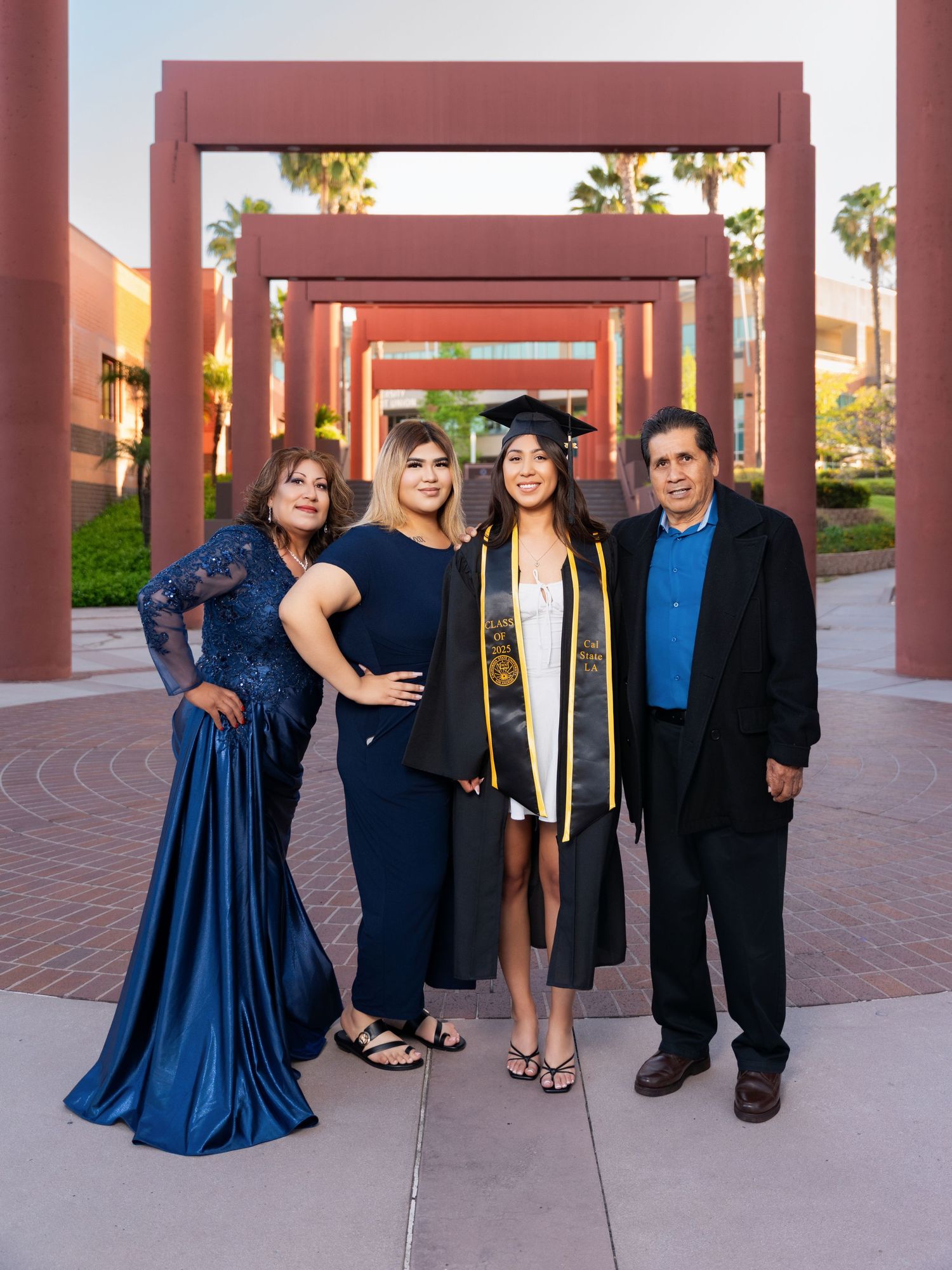 A group in formal attire poses together in front of a brick colonnade during sunset, with palm trees visible in background.