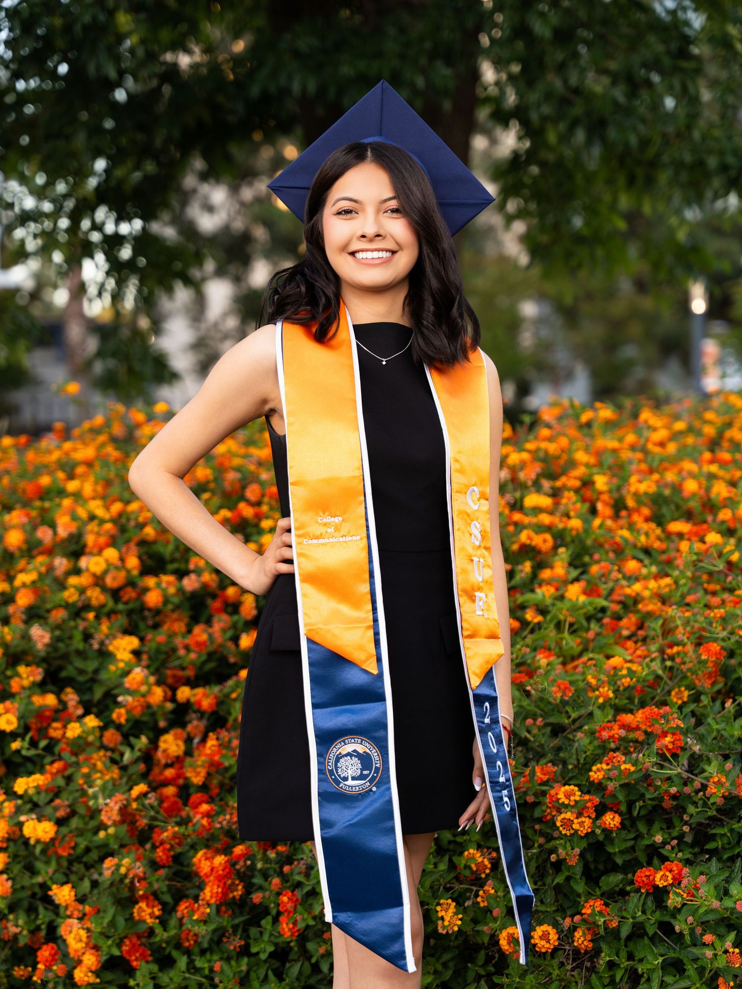 Graduate in cap and gown with gold sash poses in front of vibrant orange marigold flower garden on sunny day.