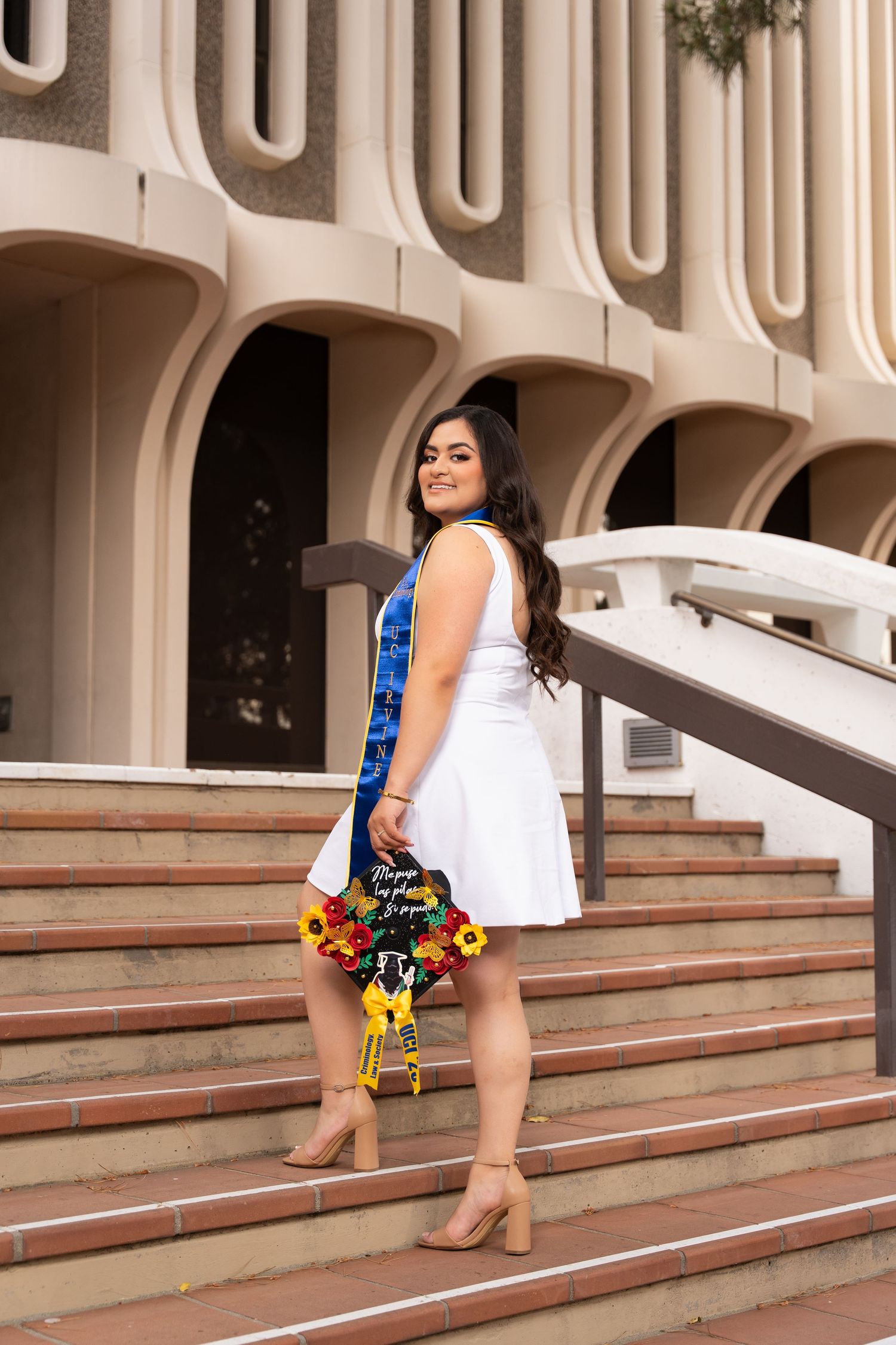 A woman in a white dress and colorful clutch poses on concrete steps with retro architectural details in the background.
