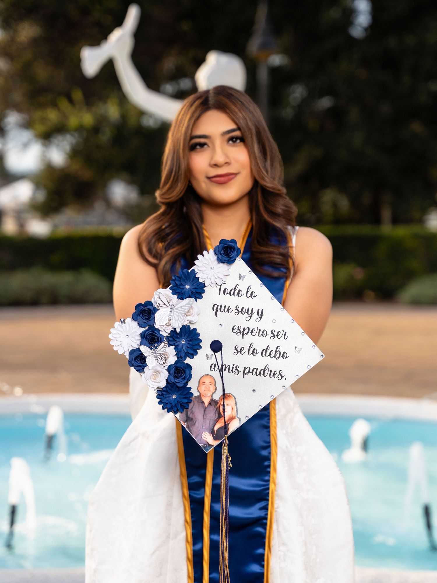Graduate in navy blue and white attire poses with decorated graduation cap near fountain pool.