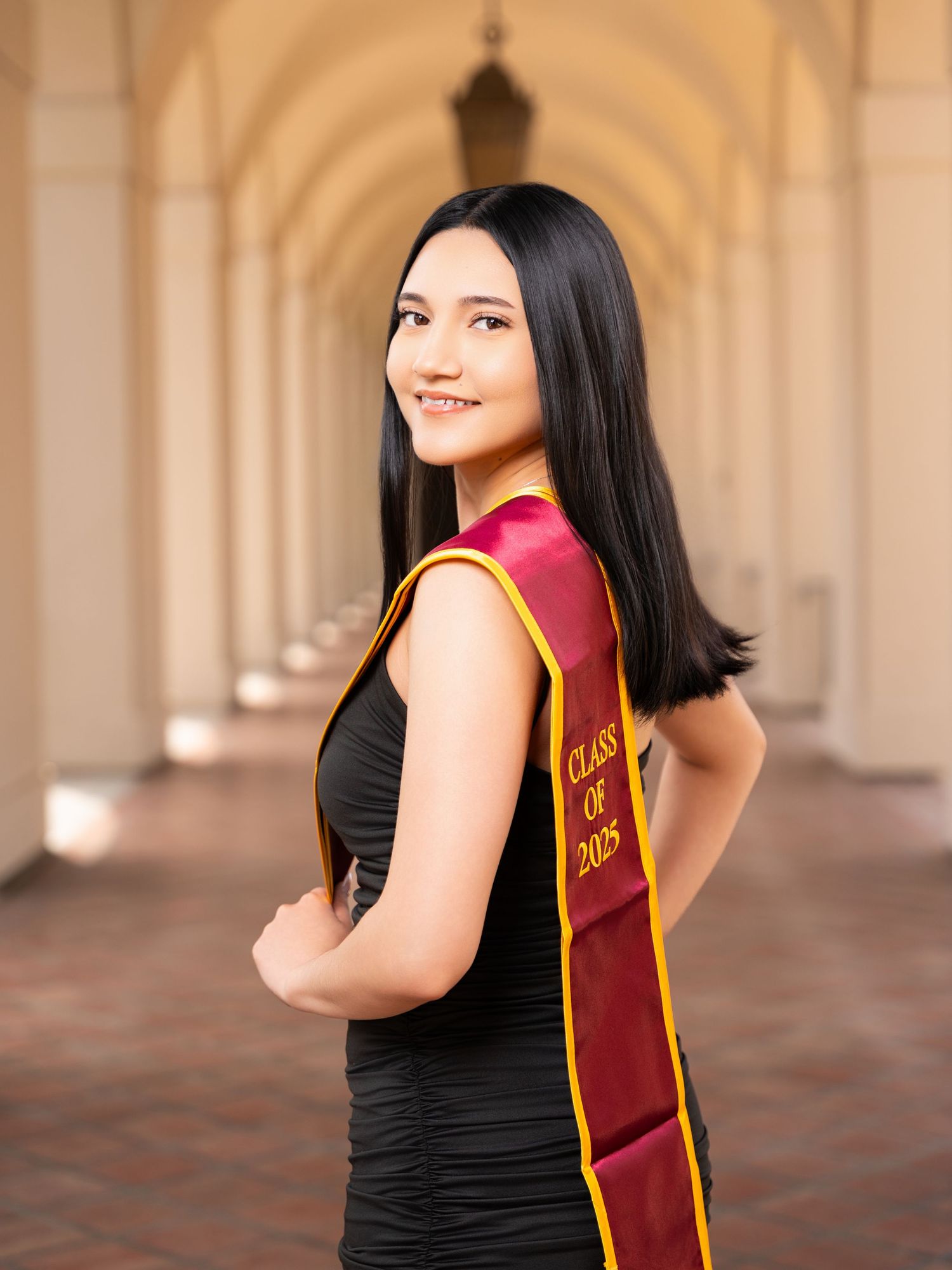 Graduate in black dress and red and gold graduation sash poses in elegant archway setting.
