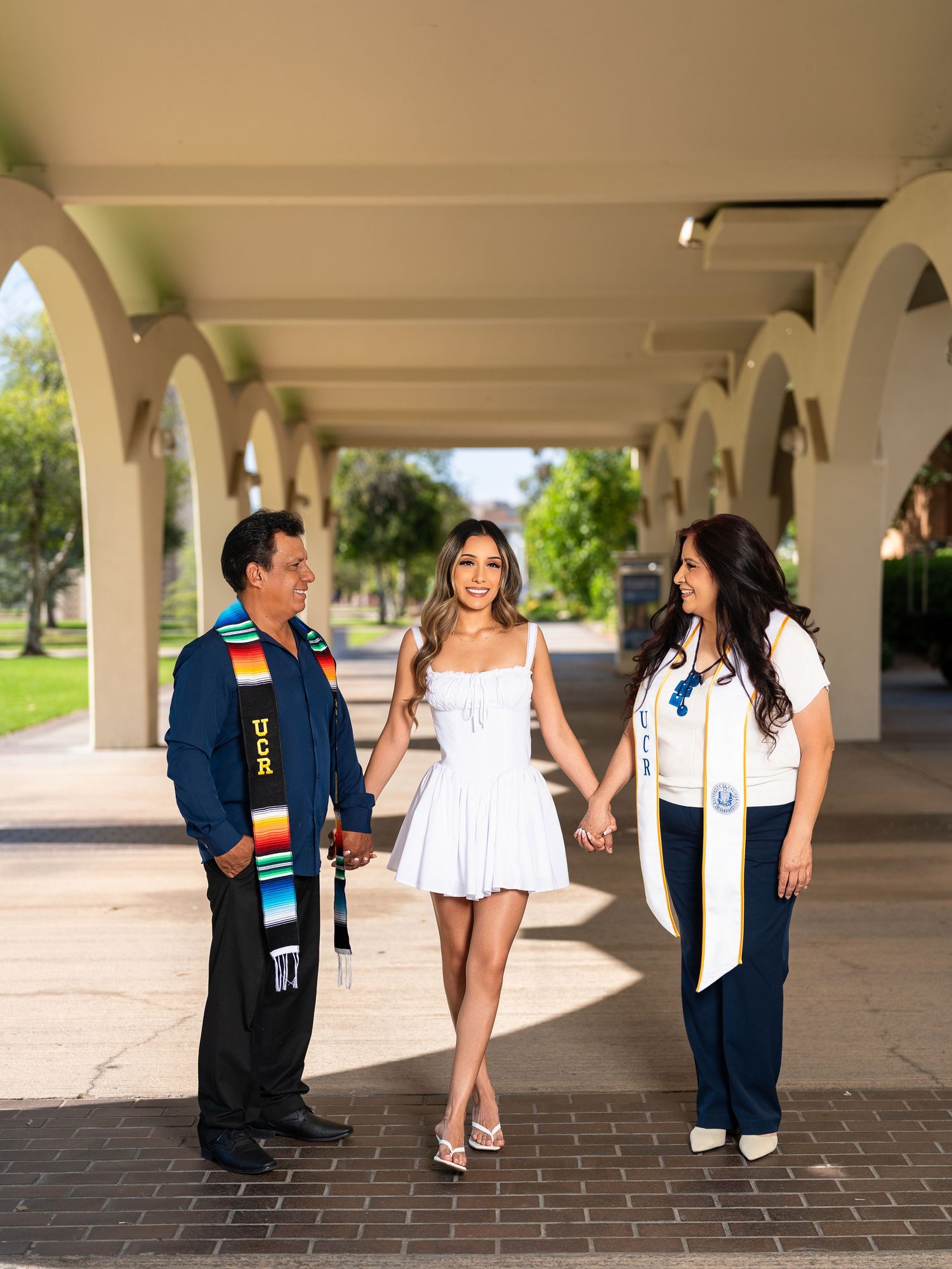 A young woman in white dress holds hands with two people wearing UCR graduation stoles under arched walkway.