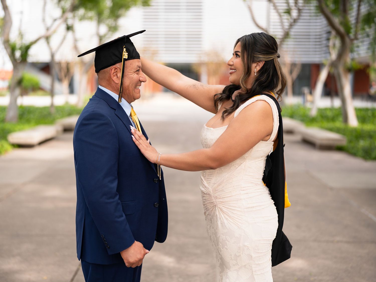 A woman in a white dress adjusts a graduation cap on a smiling man in a navy suit outdoors.
