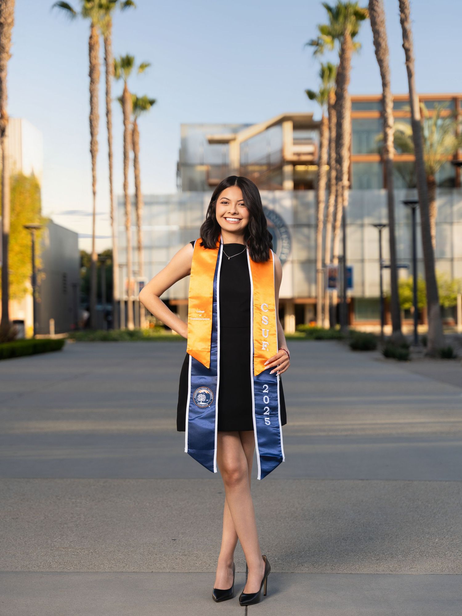 Graduate in black dress and blue sash stands confidently among palm trees at sunset on campus walkway.