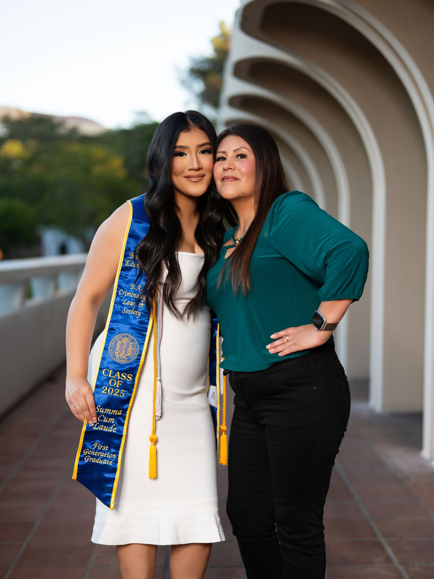 Two people sharing a joyful moment in graduation attire with blue and green clothing against an arched walkway background.