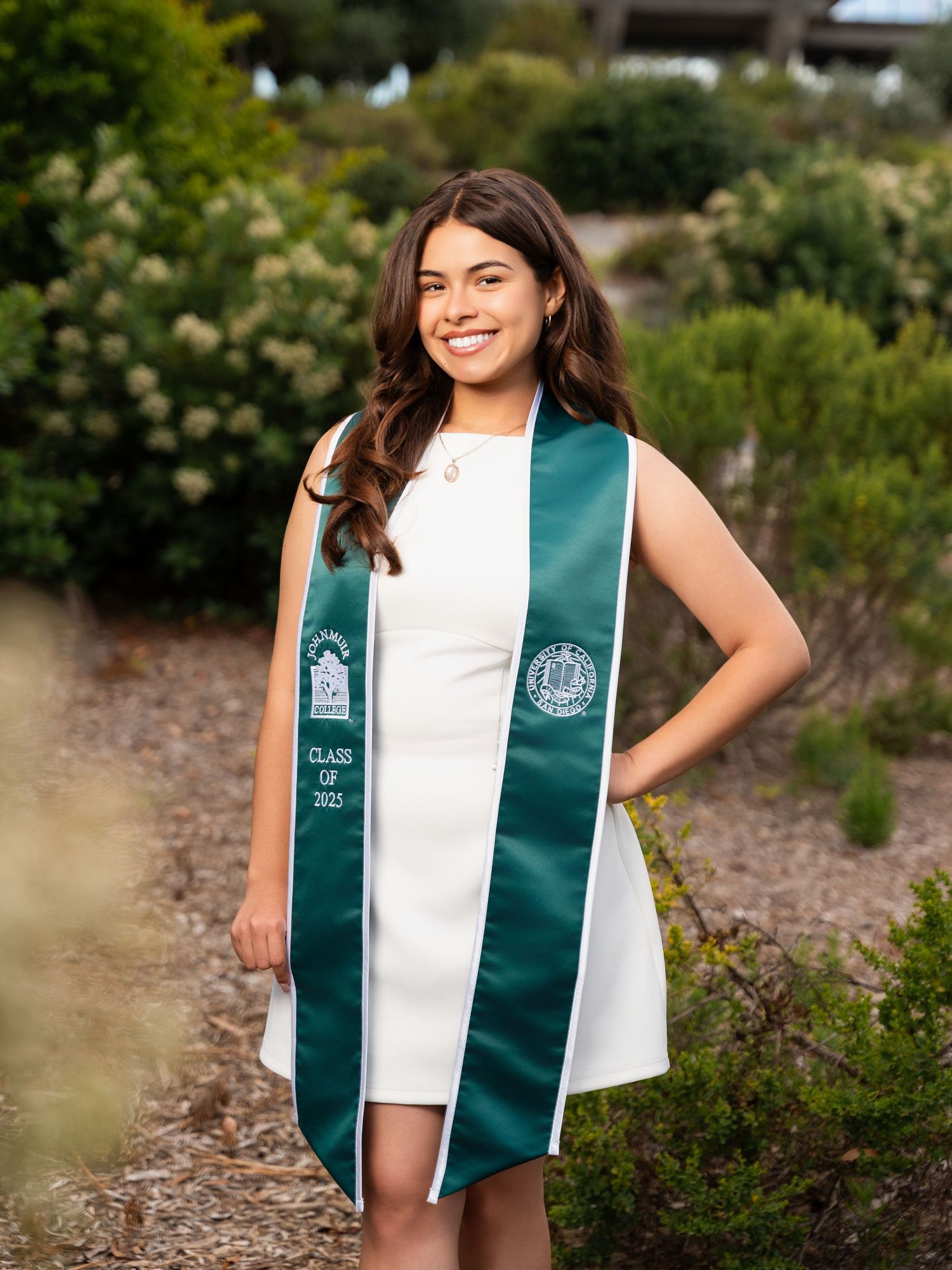 Graduate wearing a green and white graduation stole poses outdoors in a garden setting for a celebratory photo shoot.