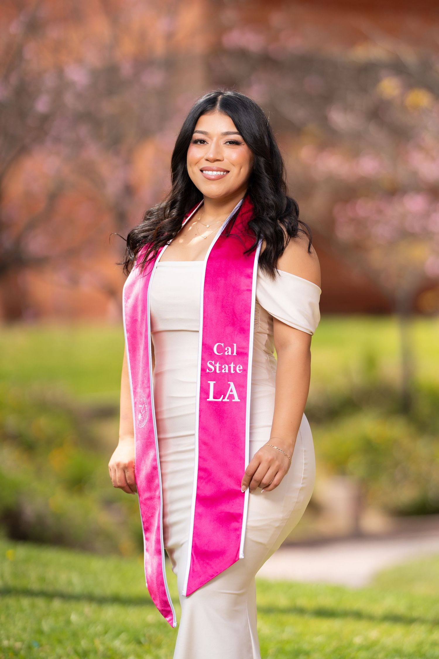 Graduate wearing white dress and pink graduation stole poses outdoors during spring with cherry blossoms in background.