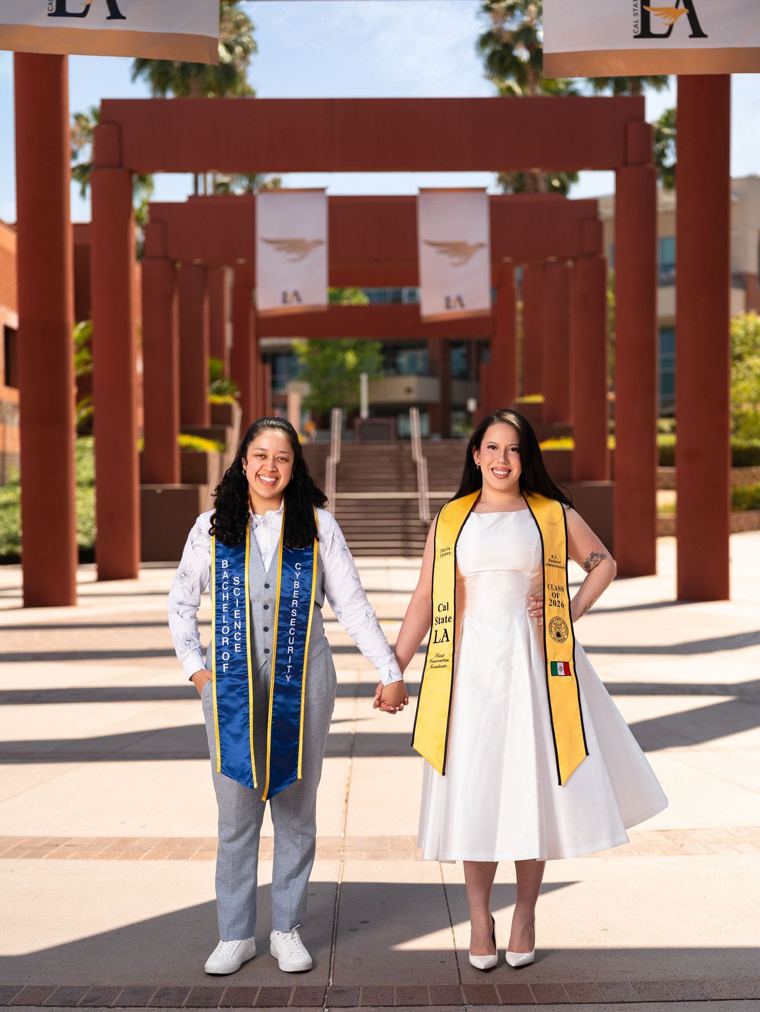 Two female graduates in academic stoles hold hands outdoors at Cal State LA campus during commencement celebration.