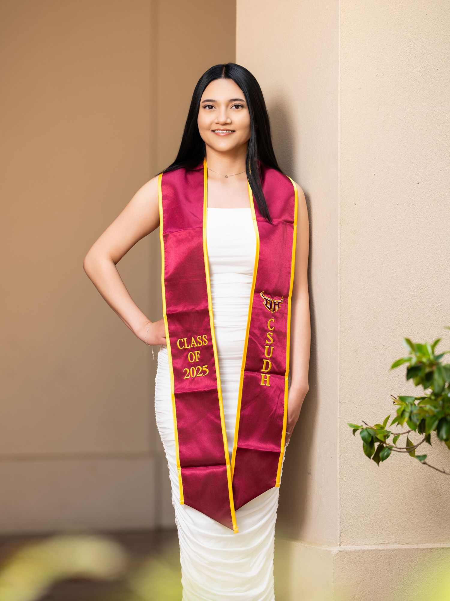 Graduate in white dress and maroon sash poses for graduation photos against neutral background.