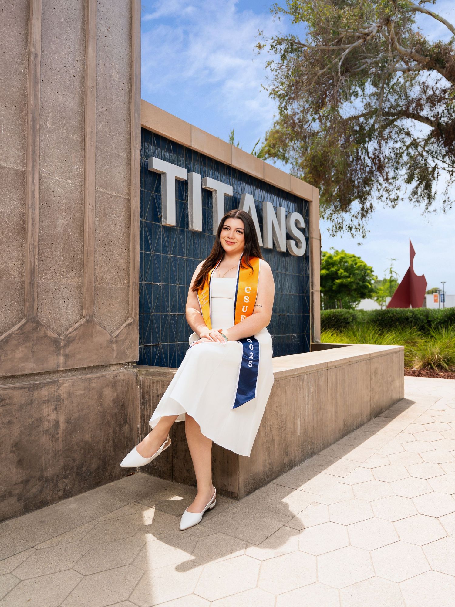 A student in white dress and orange cardigan poses for graduation photos against a concrete wall on campus.