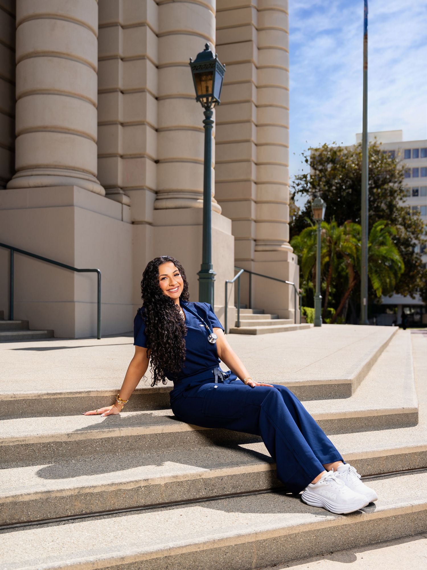 A person in a navy blue dress and white sneakers poses playfully on stone steps of a classical building.