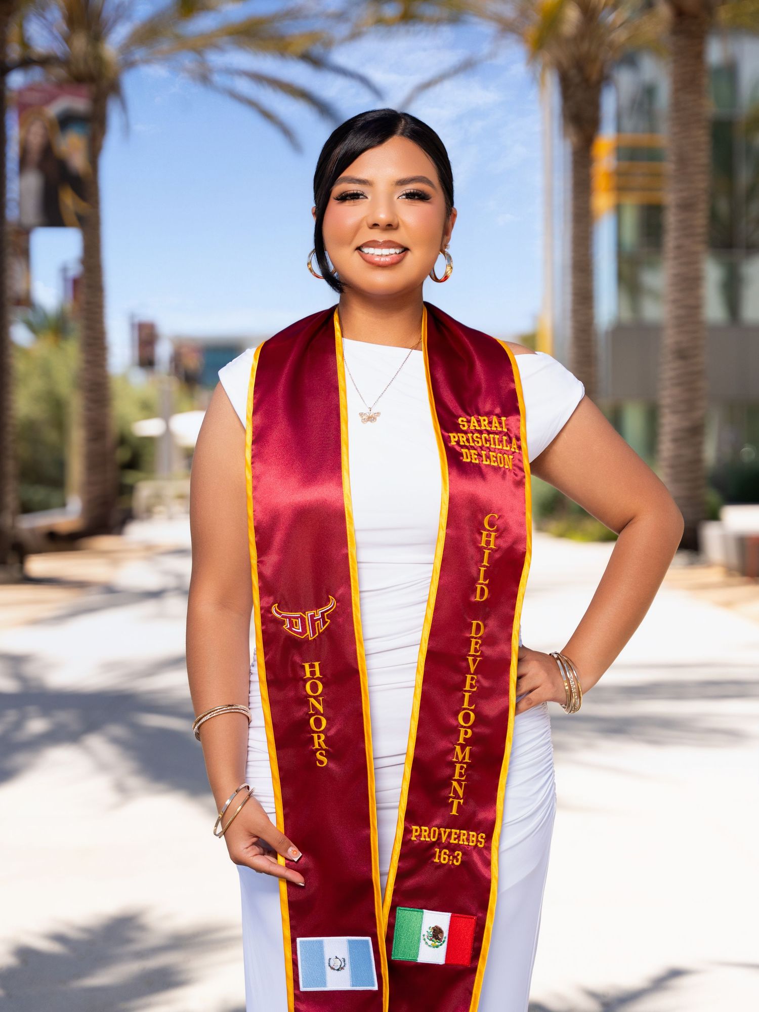Graduate wearing a burgundy and gold stole with Mexican flag emblem poses outdoors on sunny campus with palm trees.