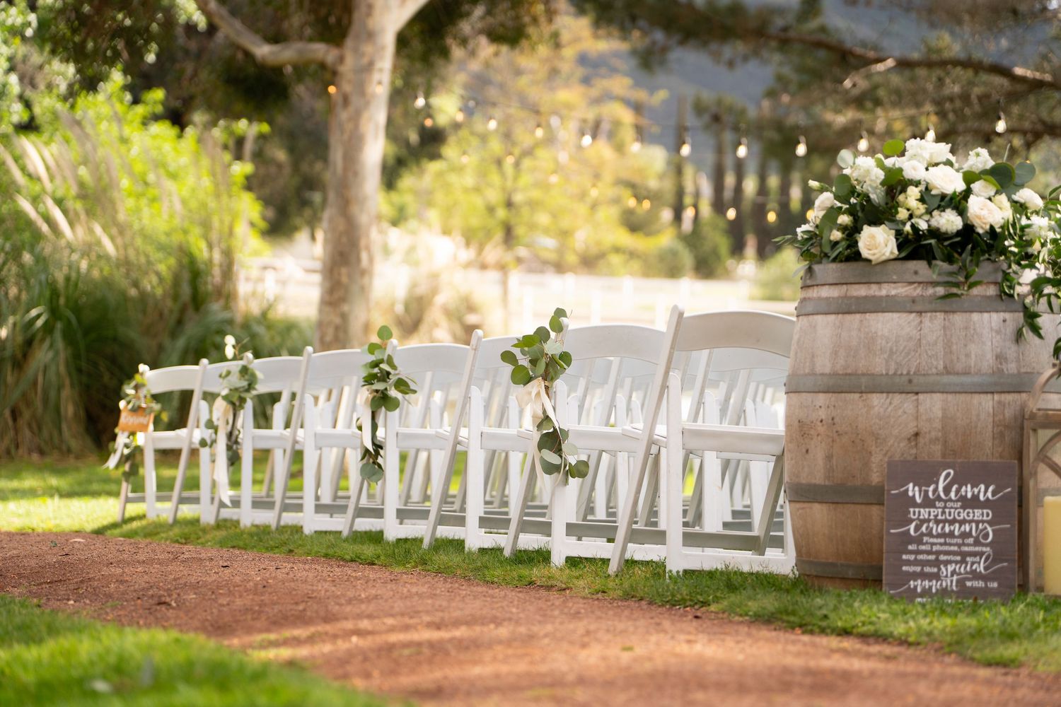 White ceremony chairs arranged outdoors with rustic barrel and welcome sign.