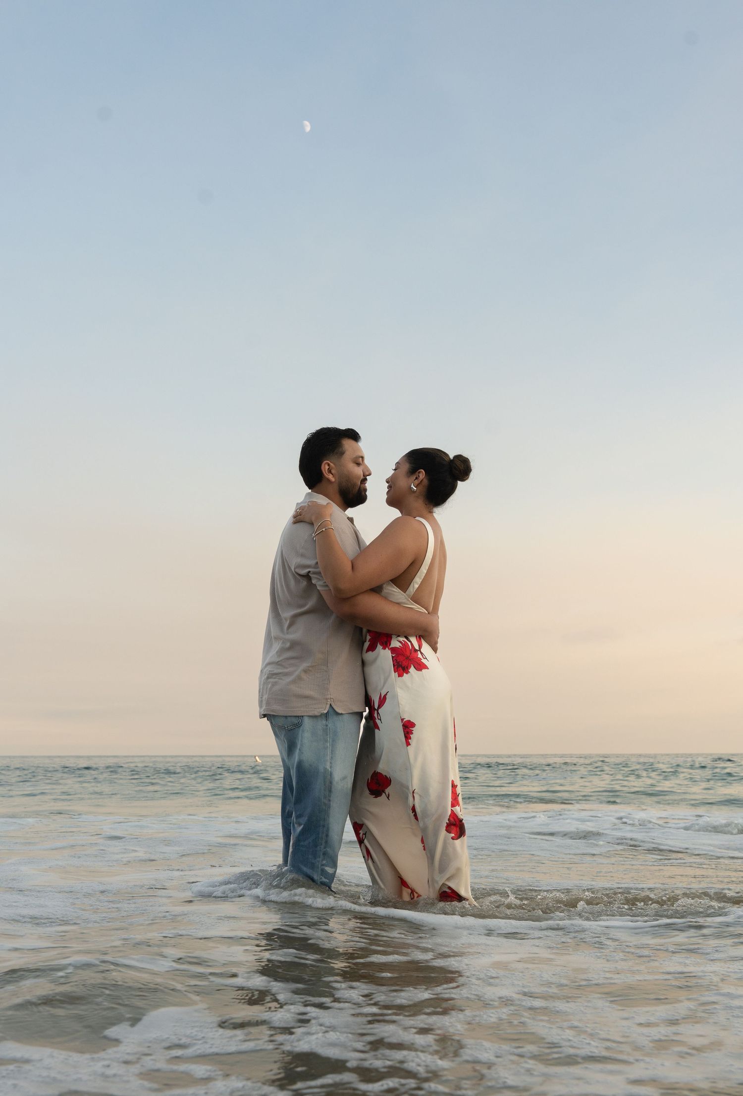 A couple embraces while standing in shallow ocean waves at sunset, with the woman wearing a floral dress.