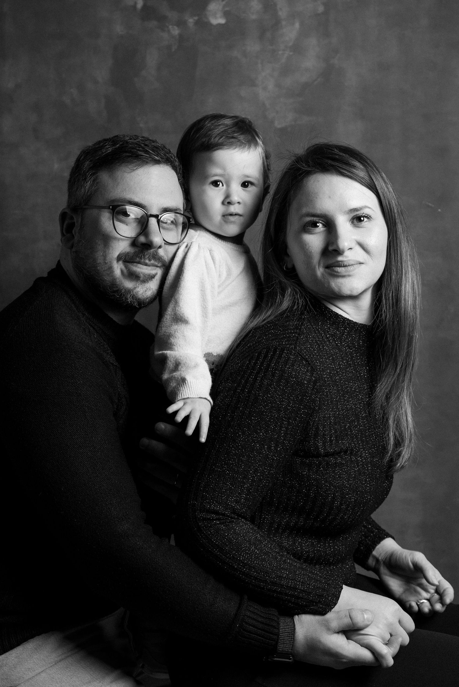 Black and white portrait of a family of three posing together against a dark background.