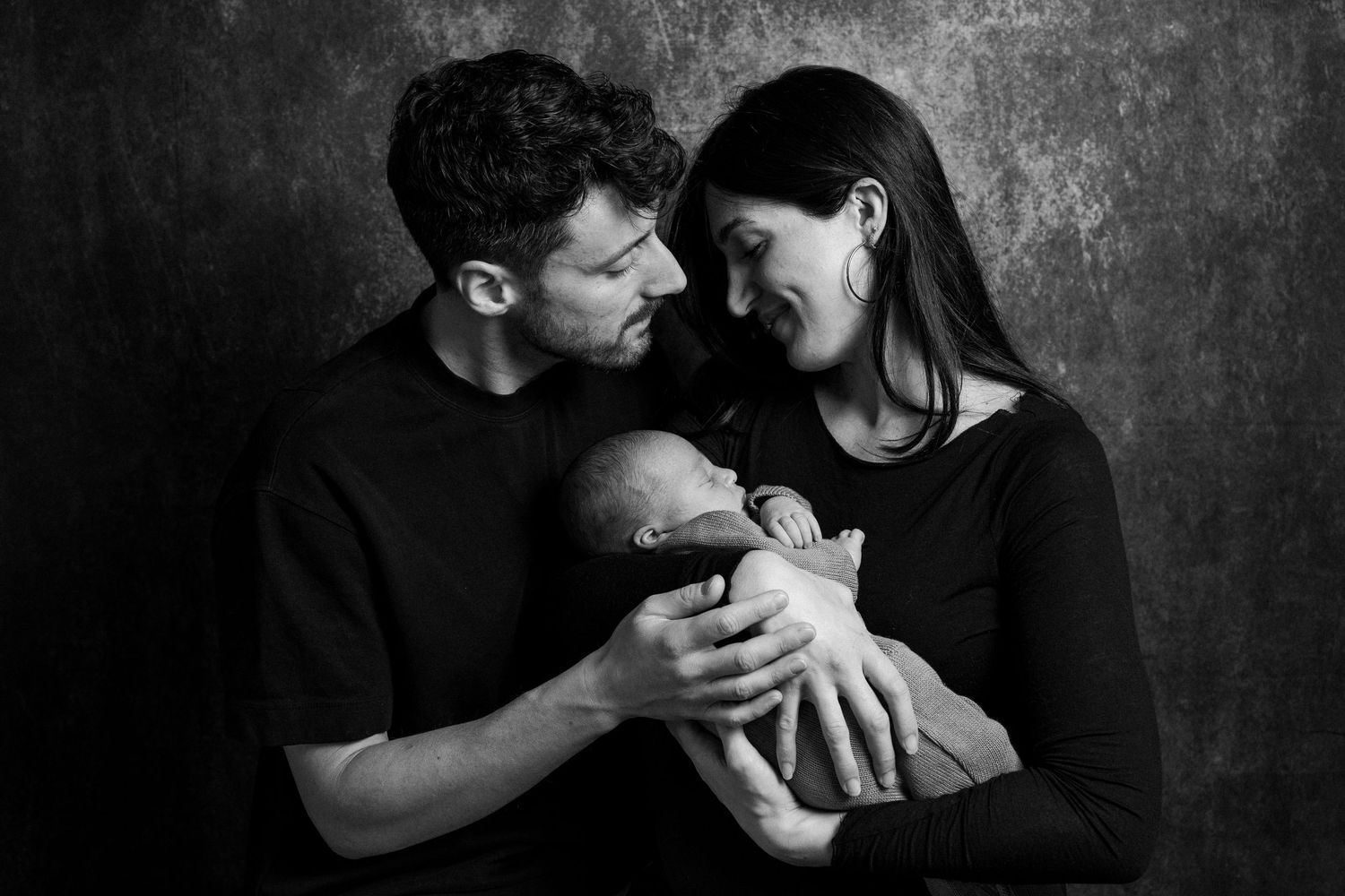 A couple in black clothing gazes lovingly at their newborn baby against a dark textured backdrop in black and white.