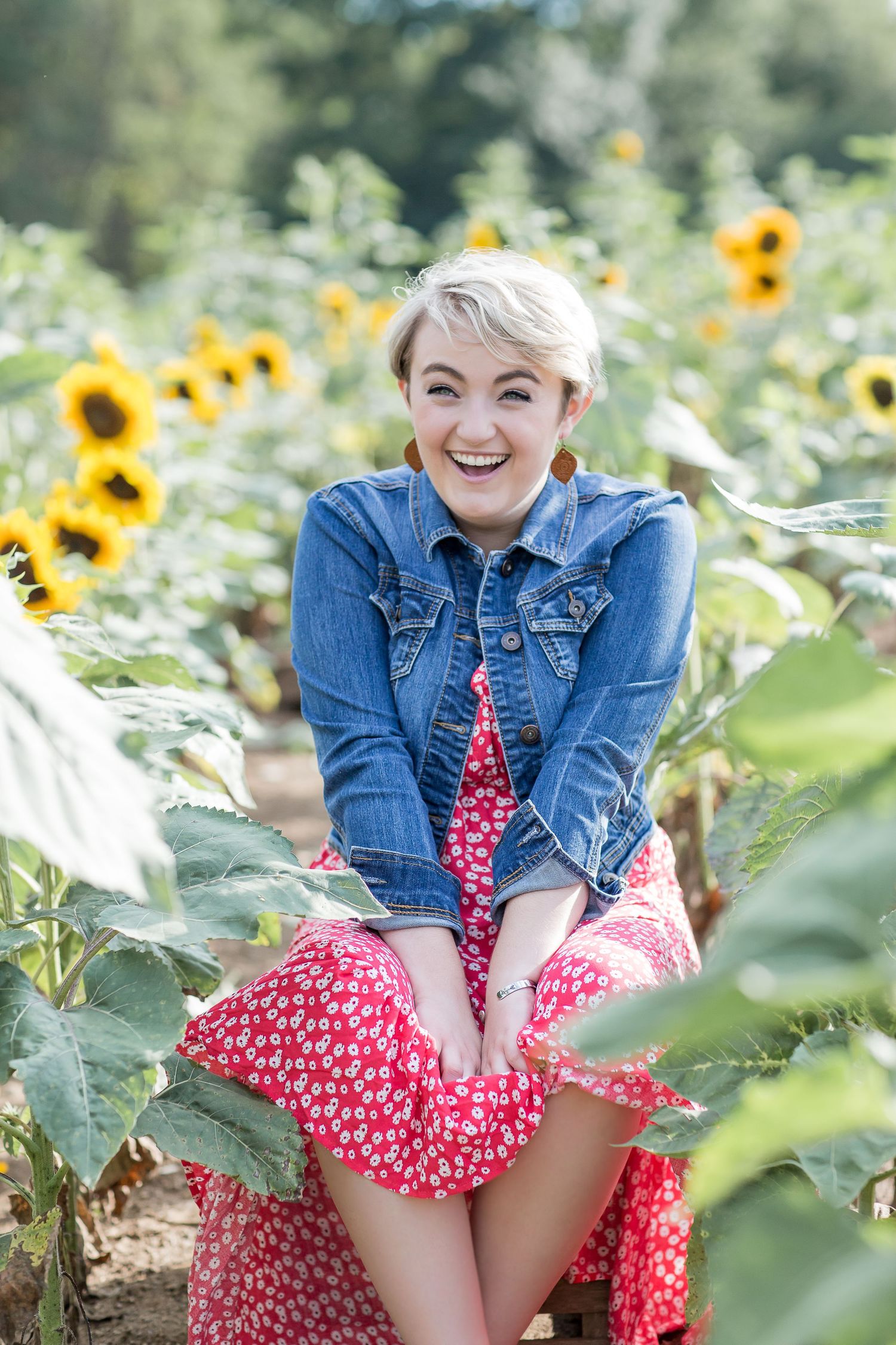 Laughing blonde woman in denim jacket and red floral dress sitting among sunflowers in a sunny field.