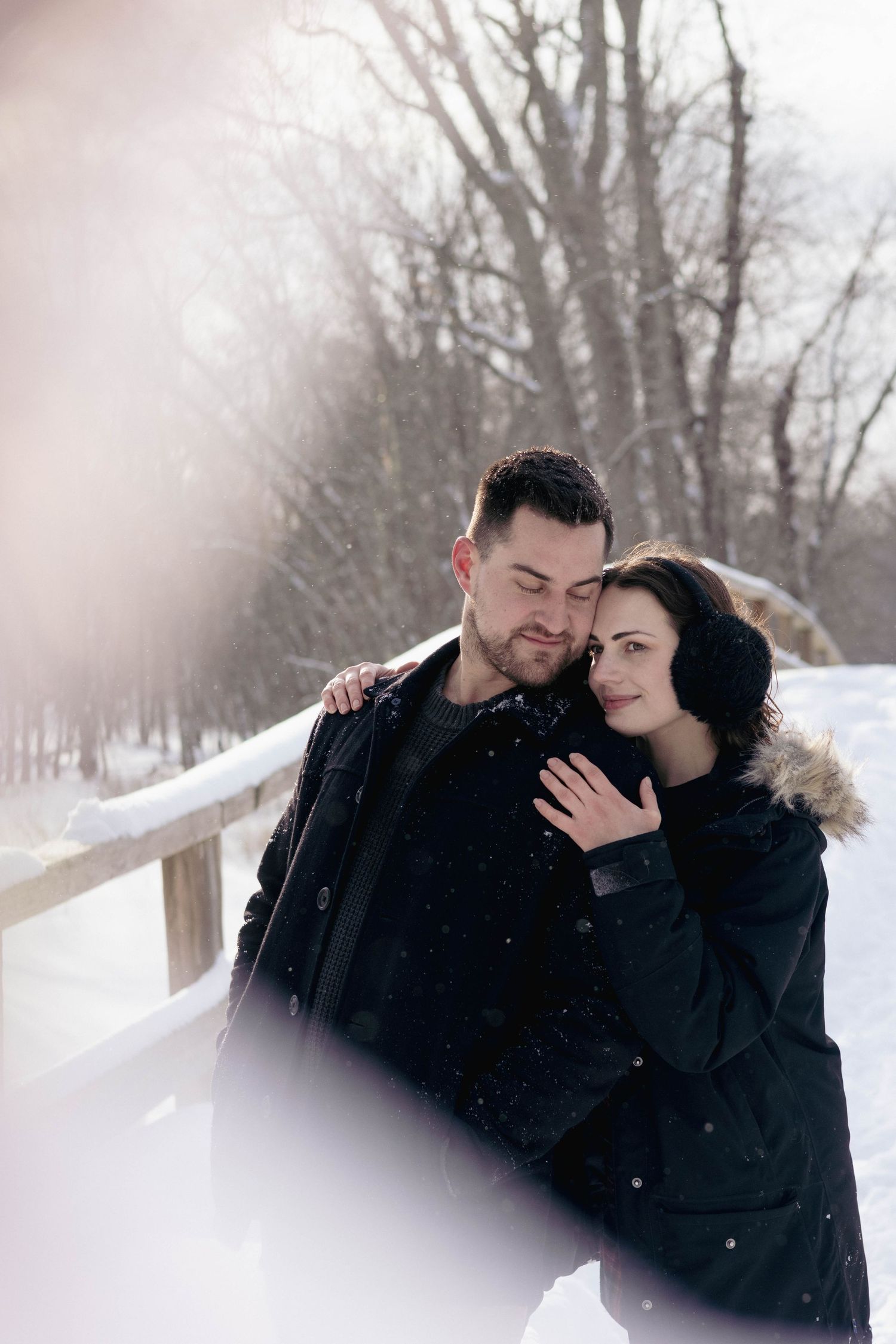 Couple embracing outdoors in falling snow, foreheads touching during a cozy winter portrait session.
