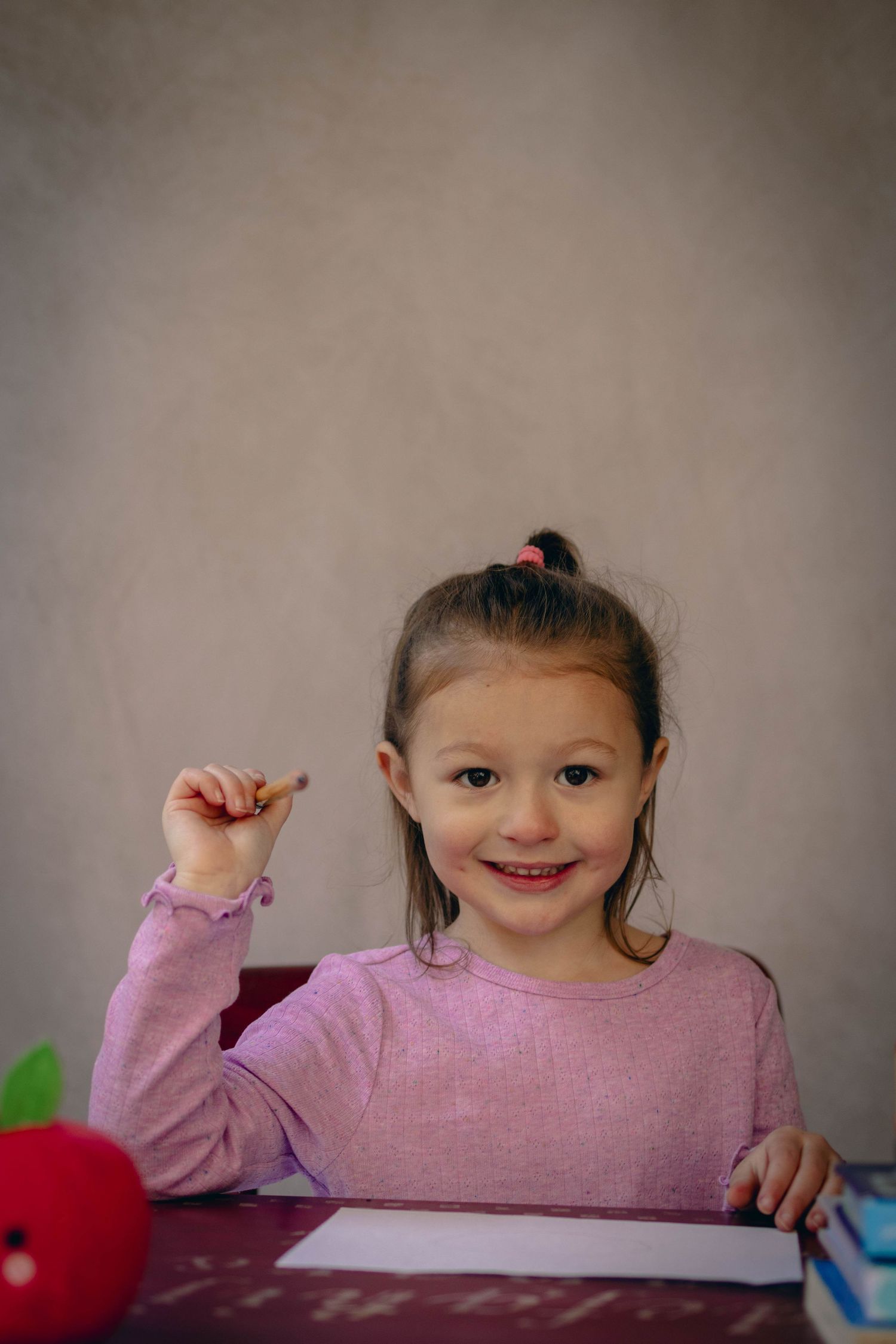 Young child sitting at a desk during a school portrait session, smiling while writing, with school‑themed props including books.