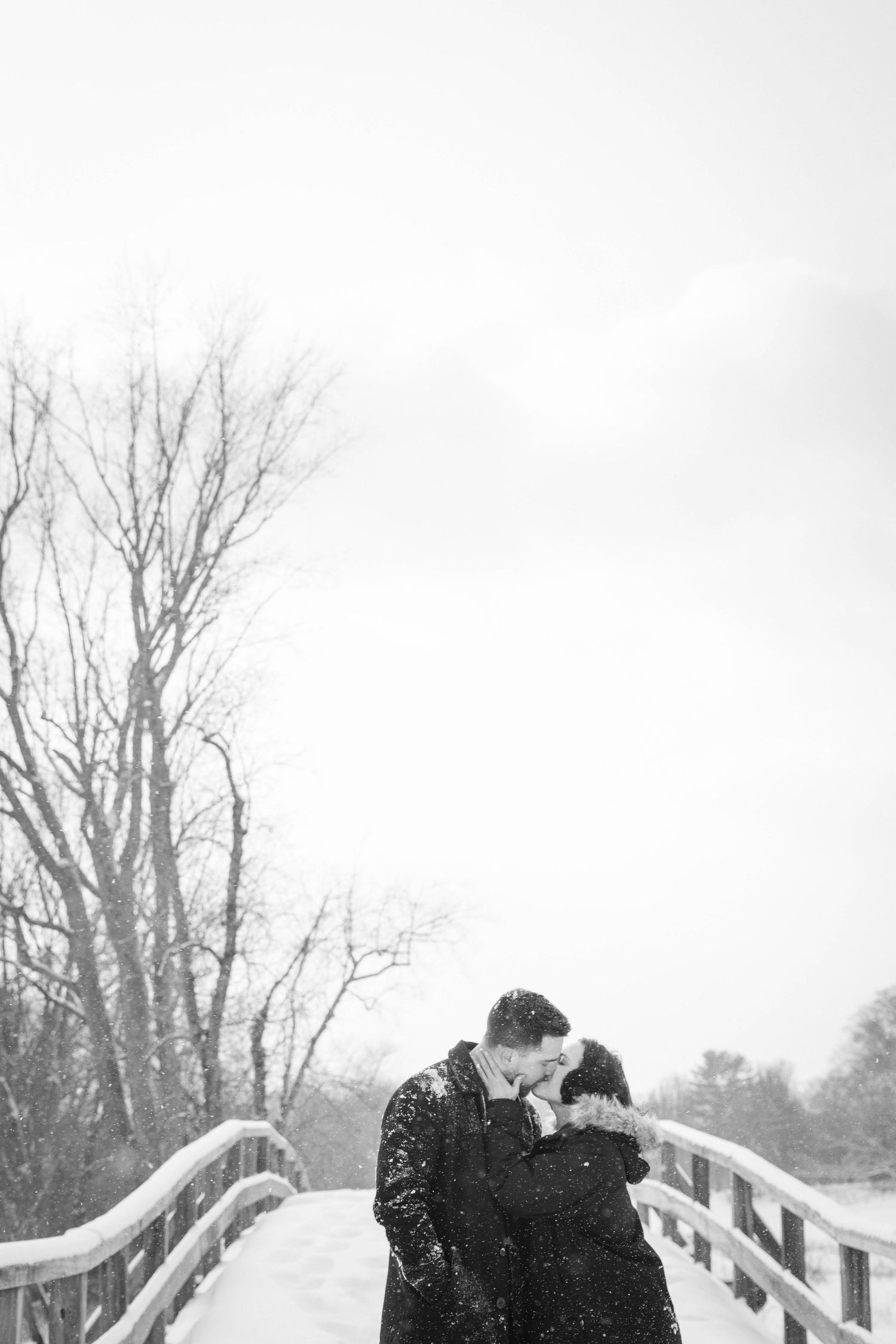 Couple embracing outdoors in falling snow, foreheads touching during a cozy winter portrait session.