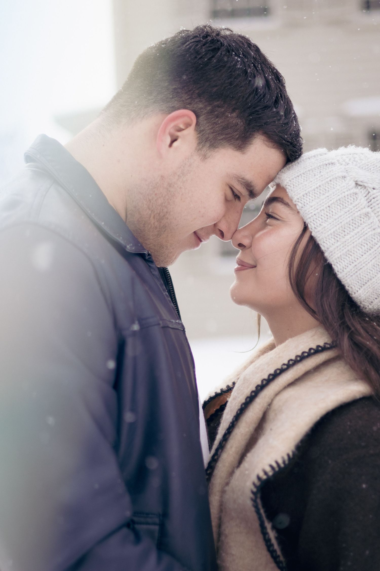 Couple embracing outdoors in falling snow, foreheads touching during a cozy winter portrait session.
