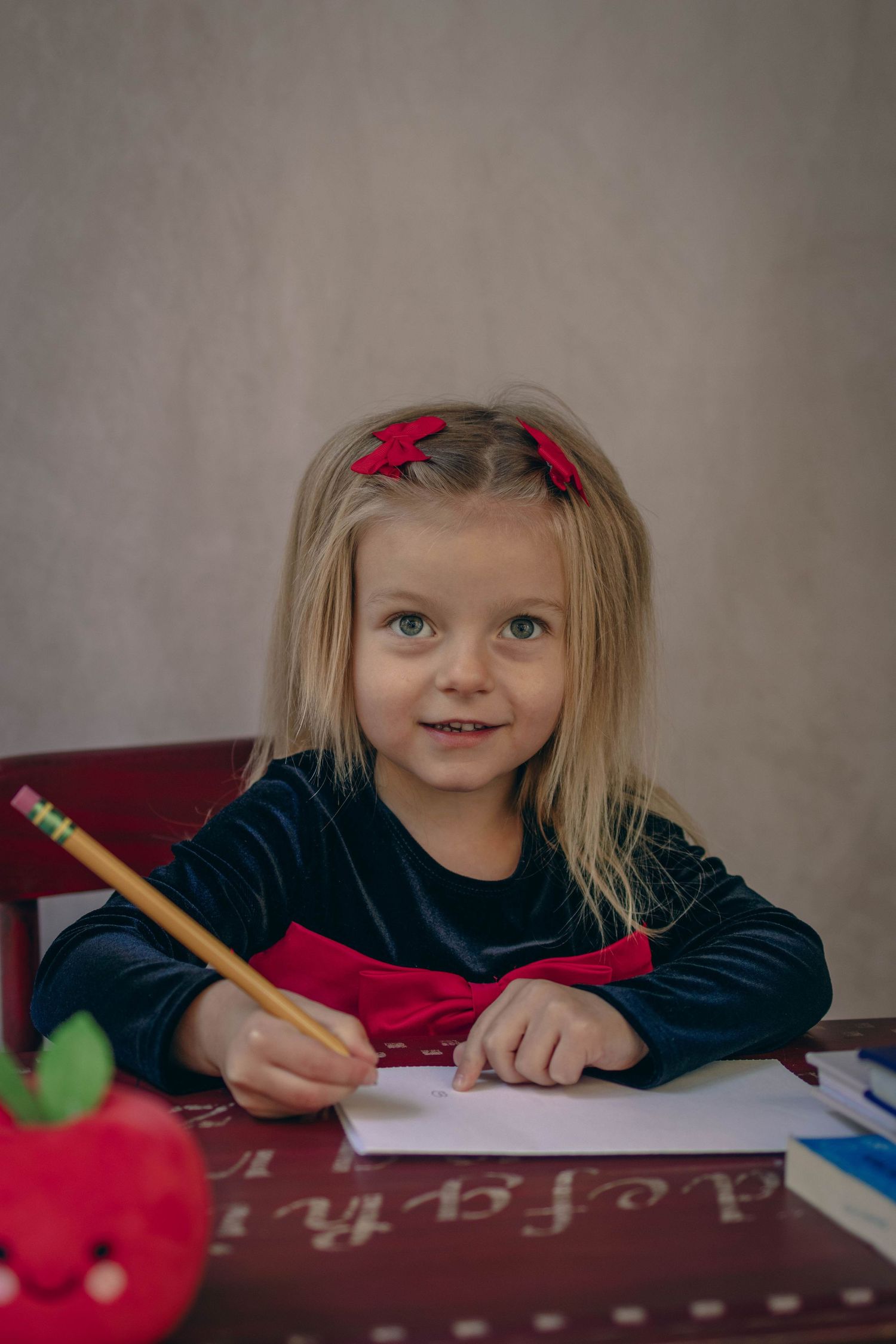 Young child sitting at a desk during a school portrait session, smiling while writing, with school‑themed props including books.