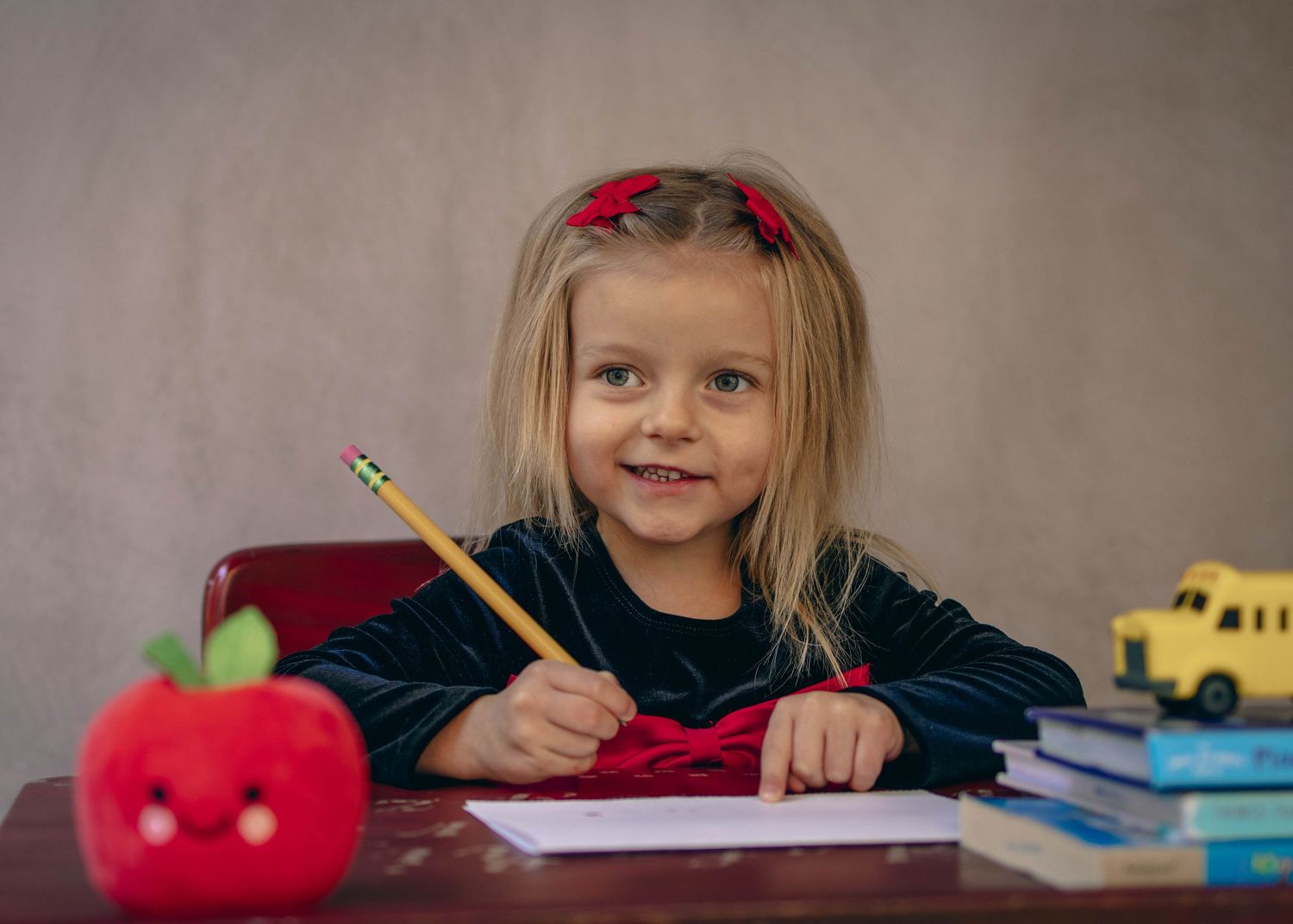 Young child sitting at a desk during a school portrait session, smiling while writing, with school‑themed props including books.