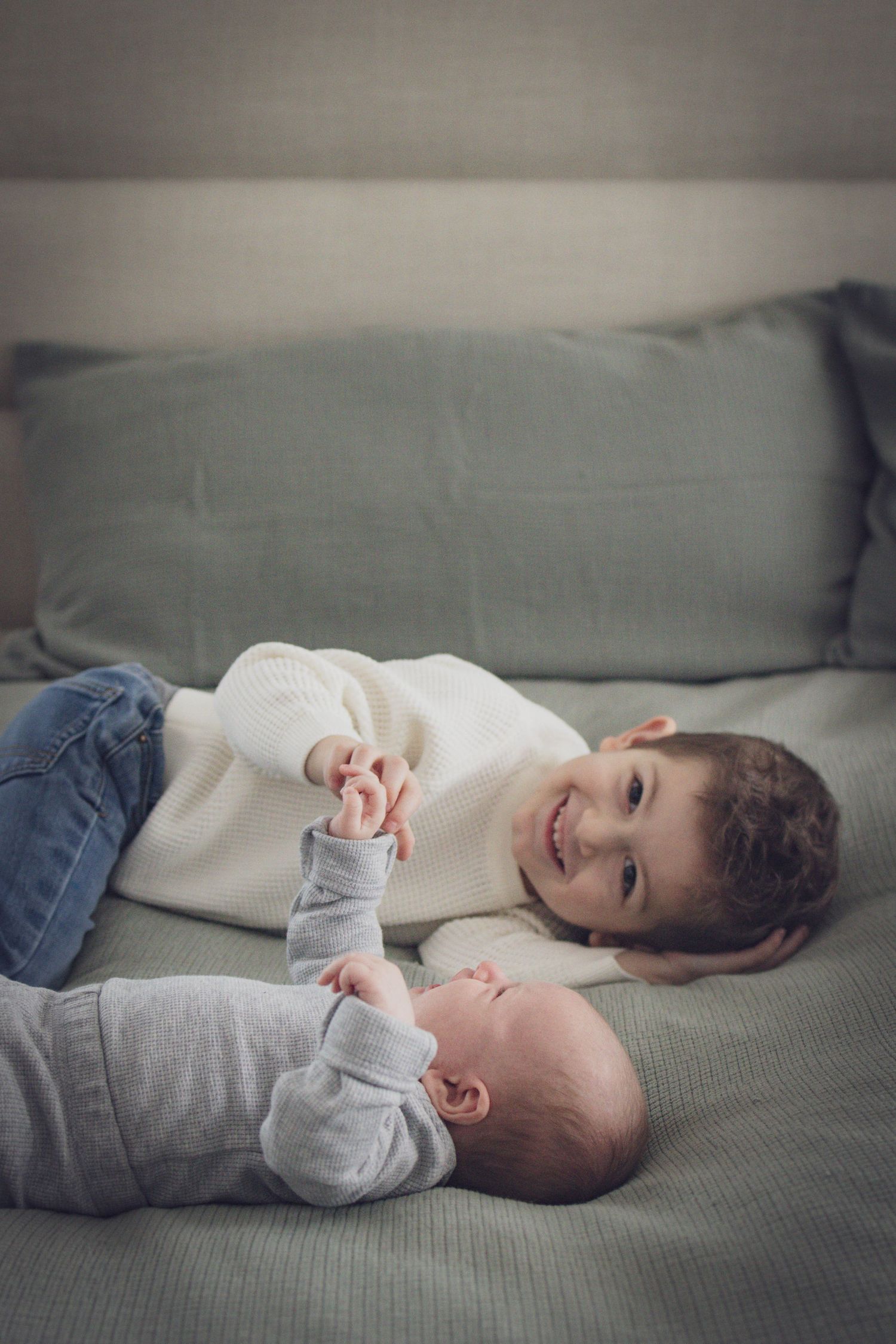 Older sibling lying on a bed and holding hands with a baby during a soft, natural family portrait session in Andover, MA.