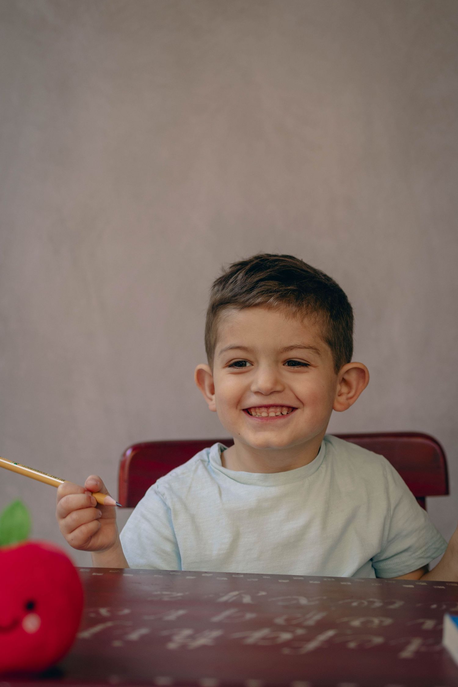 Young child sitting at a desk during a school portrait session, smiling while writing, with school‑themed props including books.