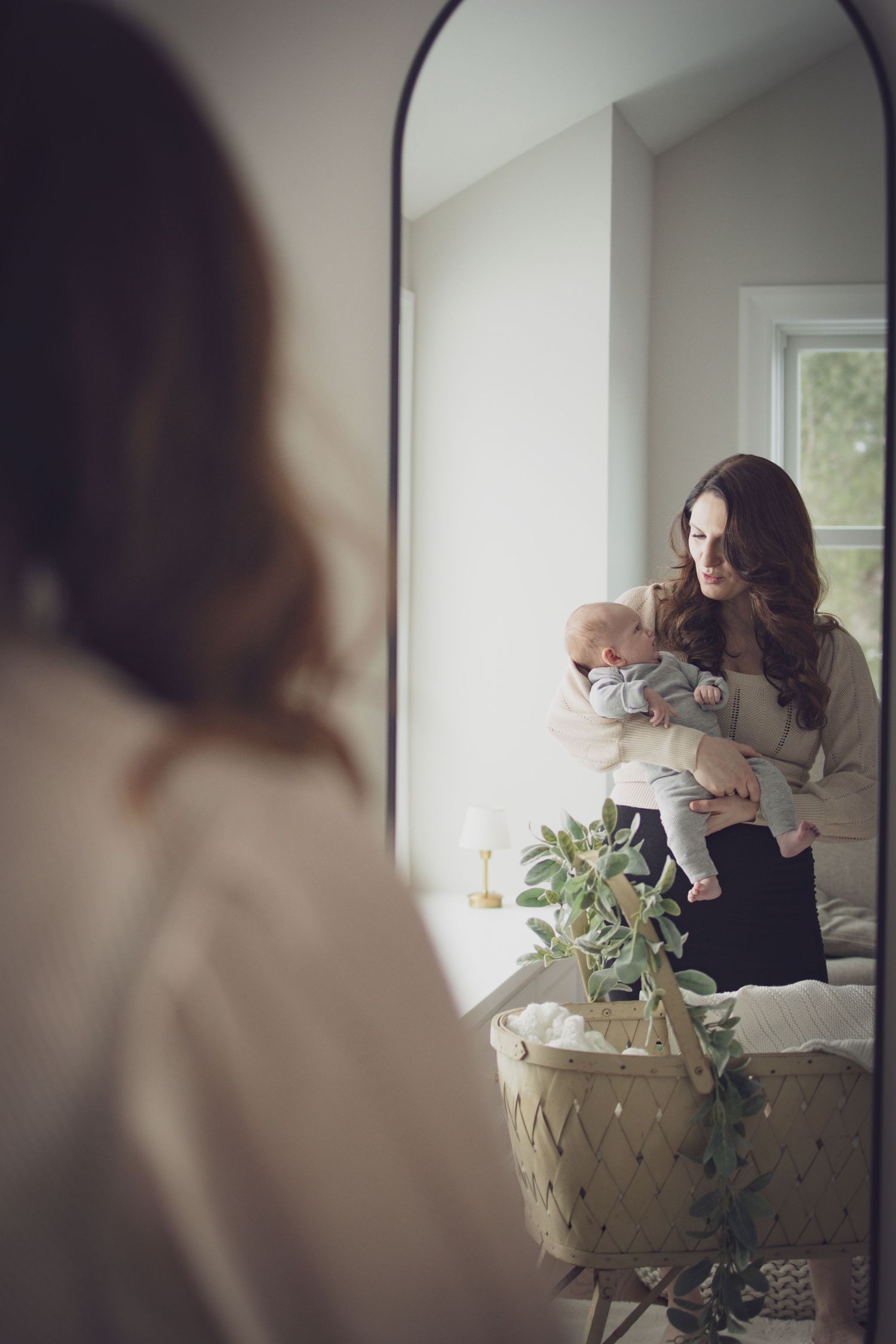 Mother holding her baby while looking into a mirror during a soft, natural indoor portrait session with warm, cozy light.