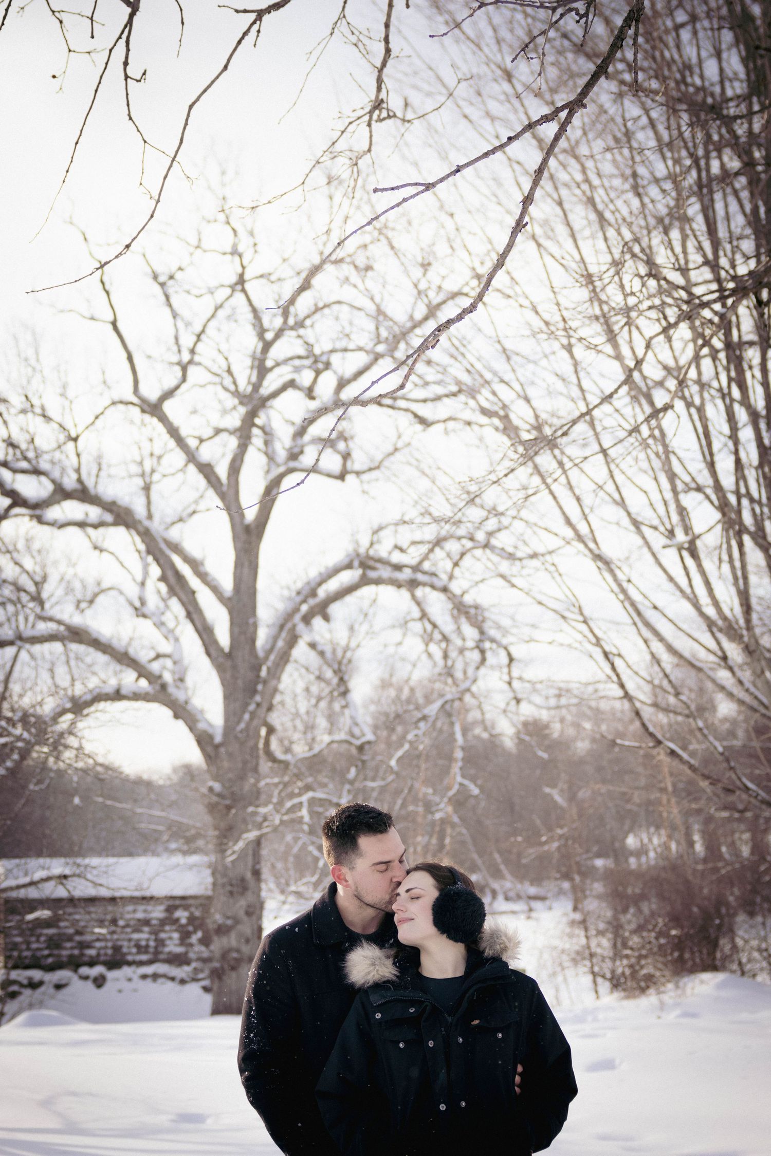 Couple embracing outdoors in falling snow, foreheads touching during a cozy winter portrait session.