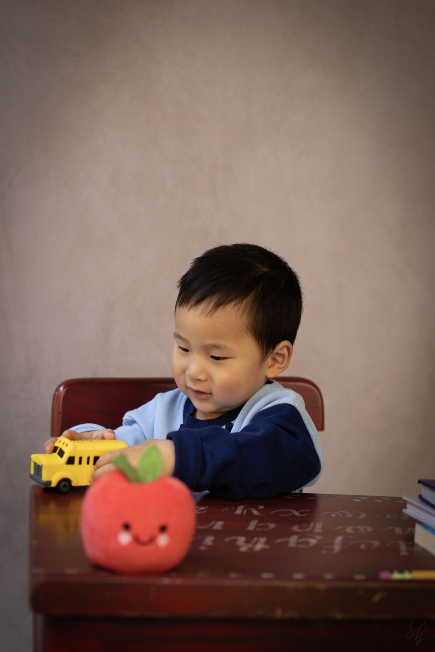 Young child sitting at a desk during a school portrait session, smiling while writing, with school‑themed props including books.