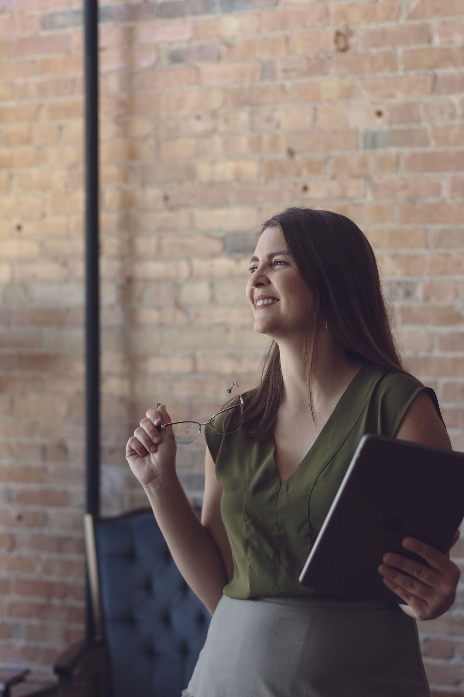 Professional branding portrait of a woman holding glasses and a tablet, standing against a brick wall with natural light.