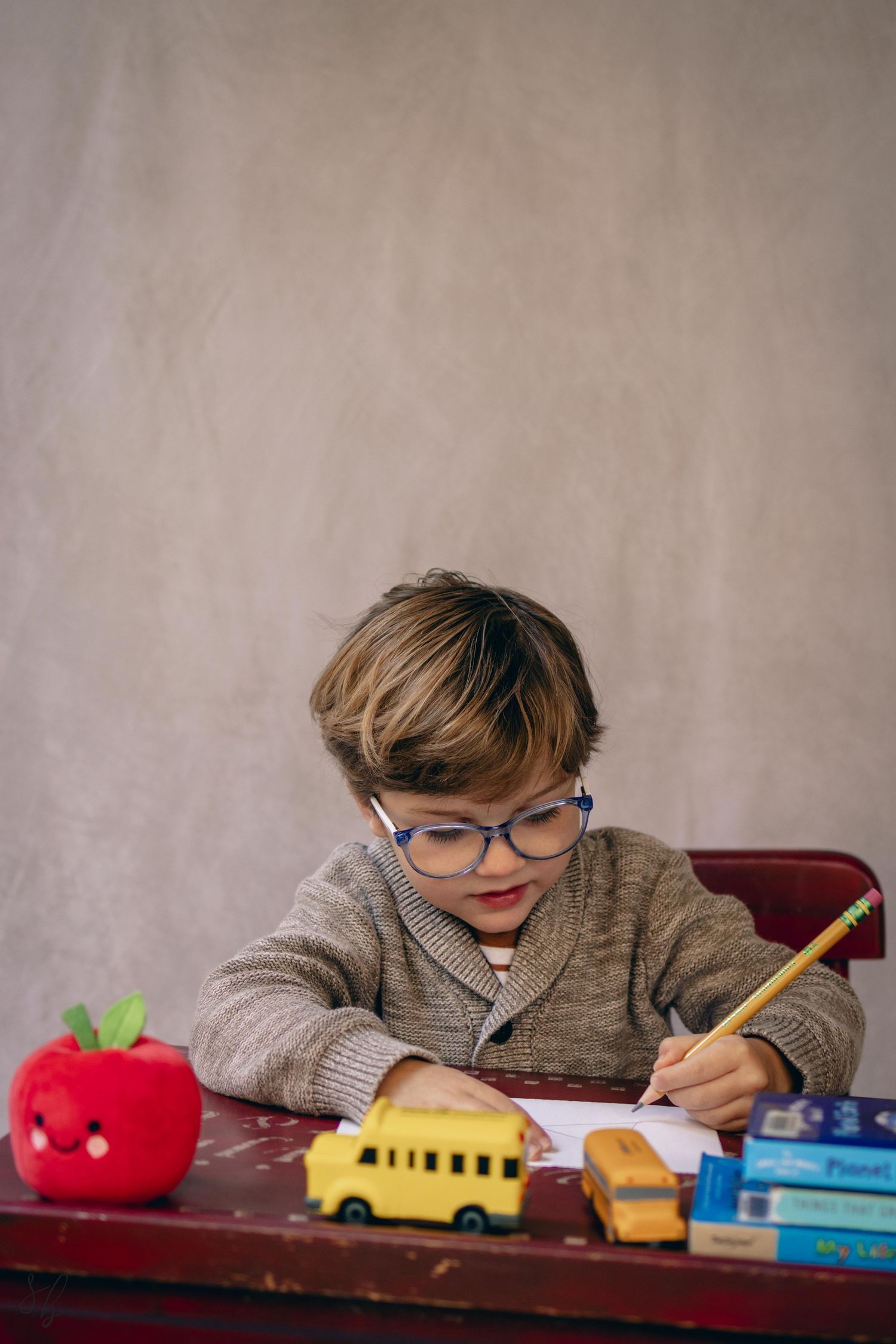 Young child sitting at a desk during a school portrait session, smiling while writing, with school‑themed props including books.