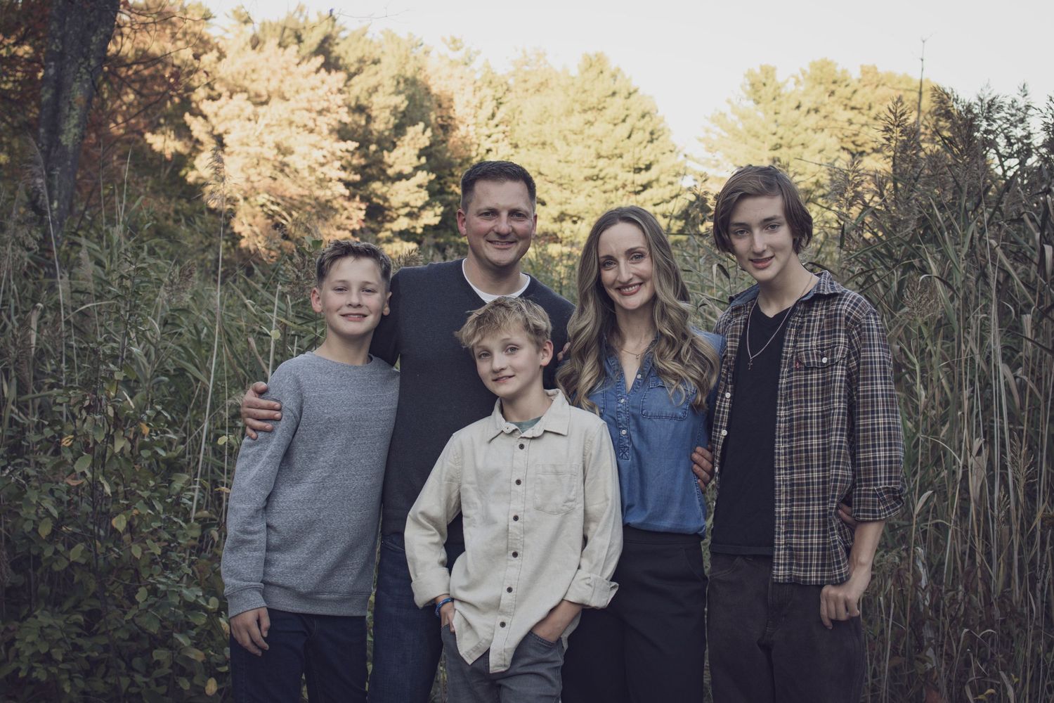 Andover family photographer capturing a fall family portrait of five people standing together in tall grass with autumn foliage.