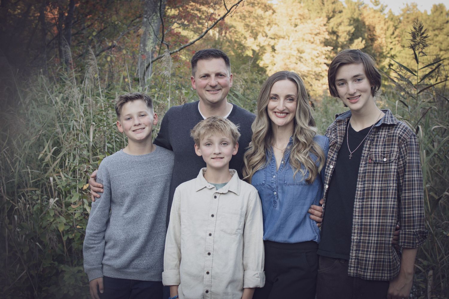 Andover family photographer capturing a fall family portrait of five people standing together in tall grass with autumn foliage.