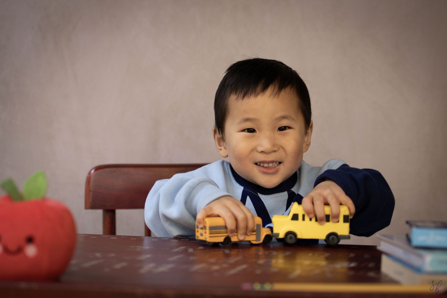 Young child sitting at a desk during a school portrait session, smiling while writing, with school‑themed props including books.