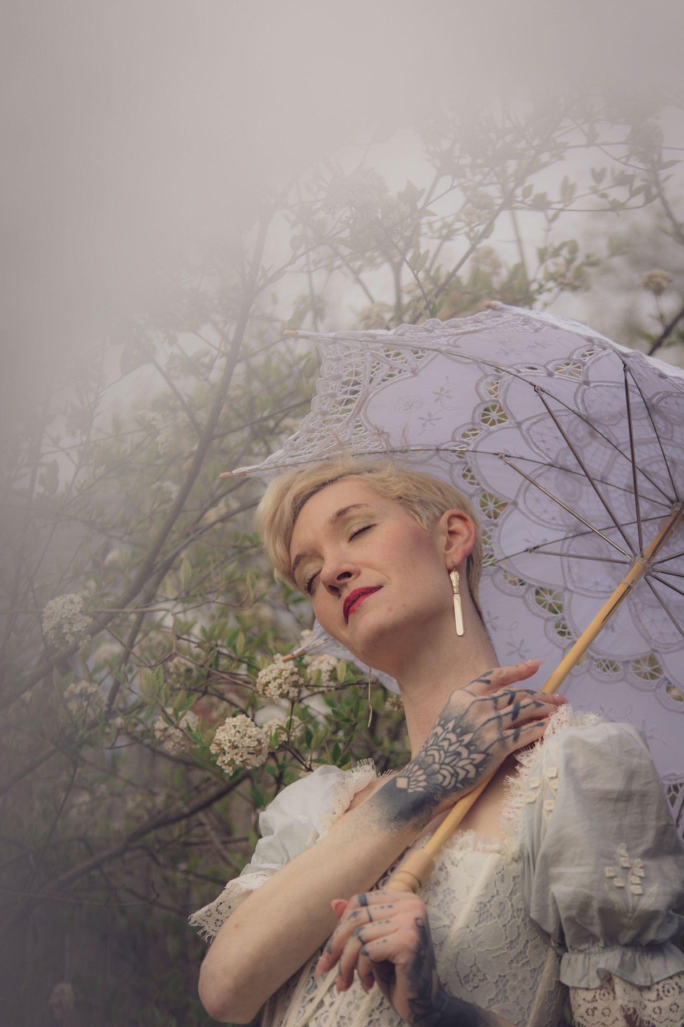 Portrait of a woman with short blonde hair holding a lace parasol among blooming spring branches, wearing a vintage white lace dress.