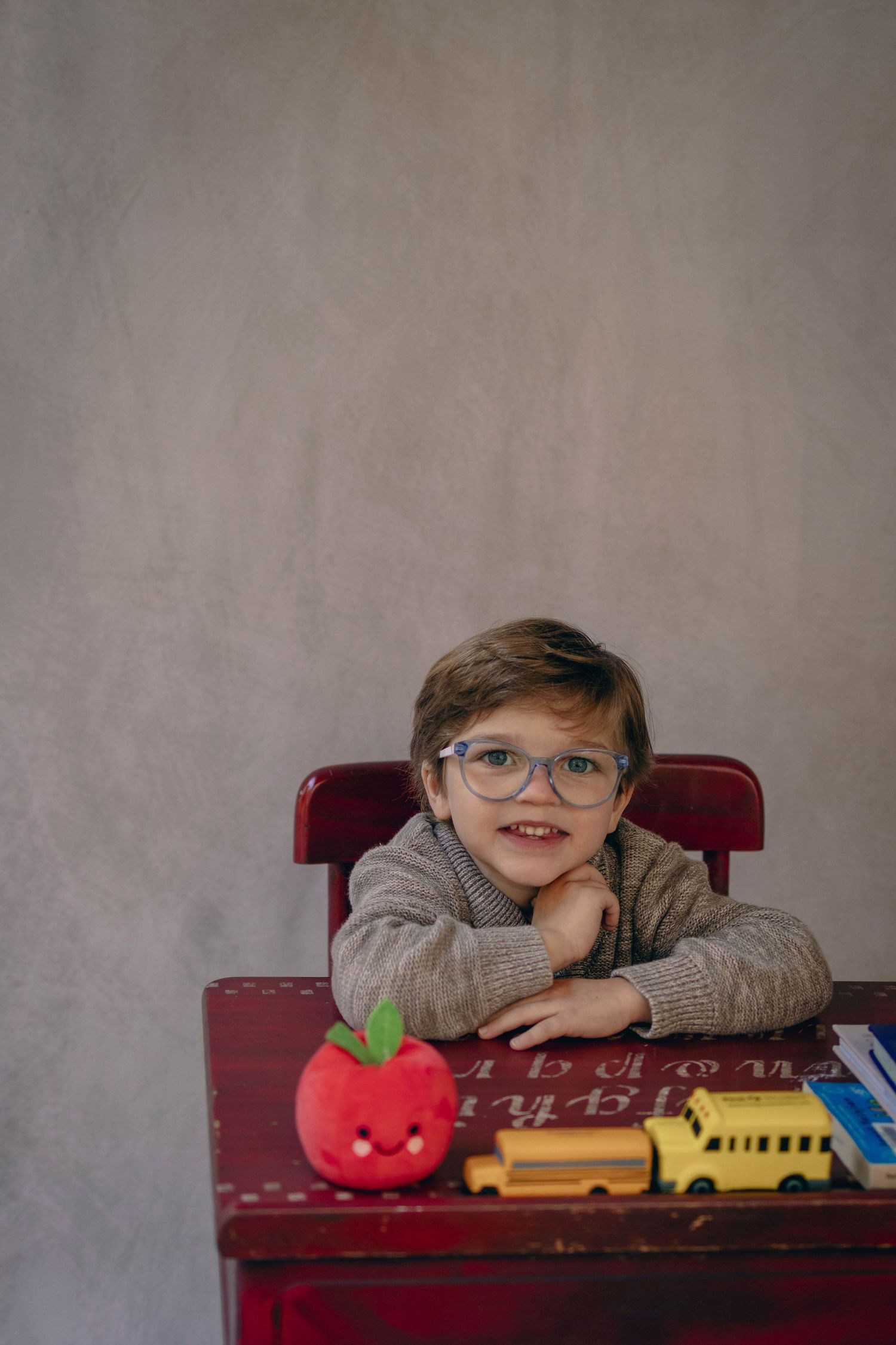 Young child sitting at a desk during a school portrait session, smiling while writing, with school‑themed props including books.