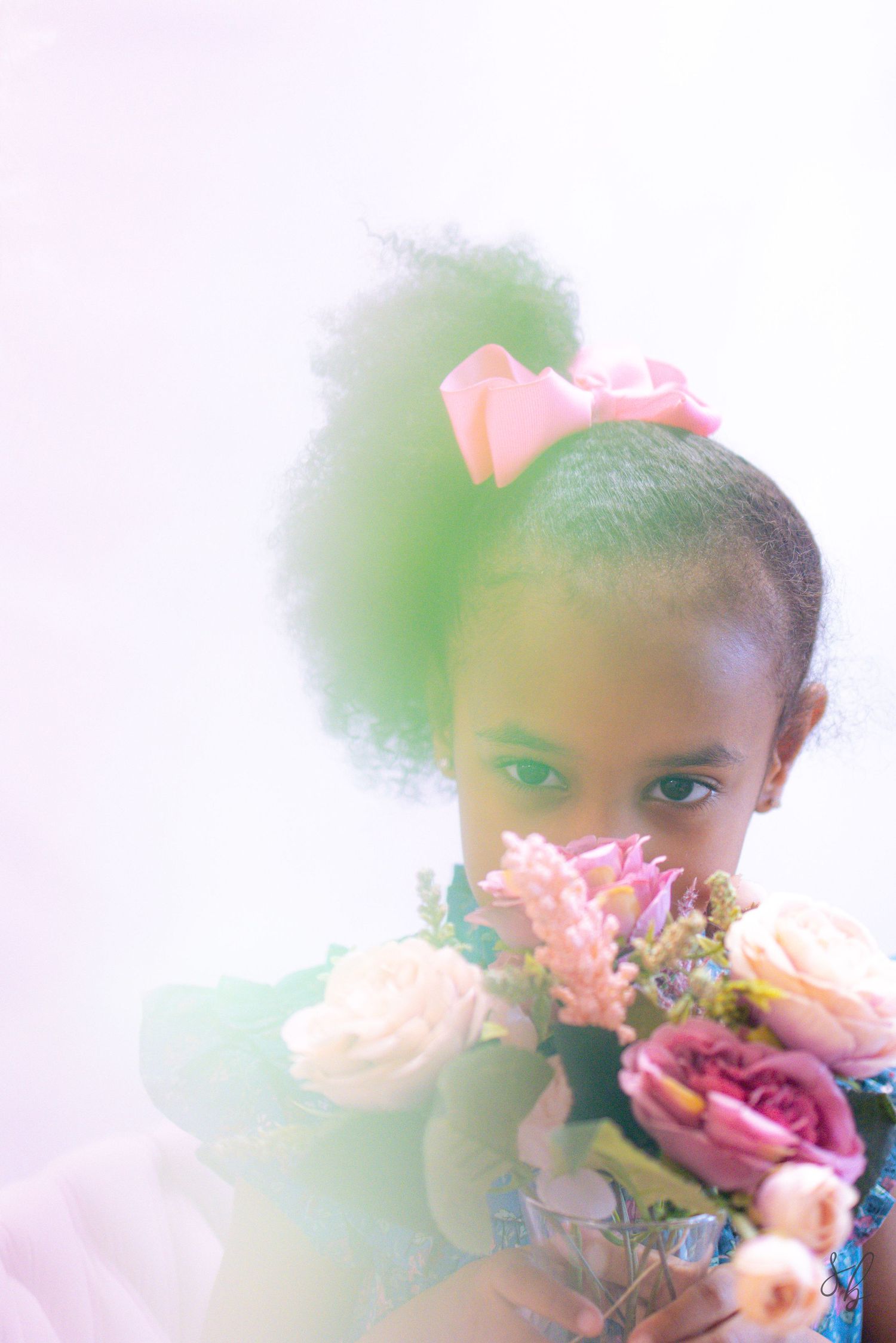 Young child holding a pastel flower bouquet during a soft, dreamy portrait session, with a large pink bow and gentle natural light.