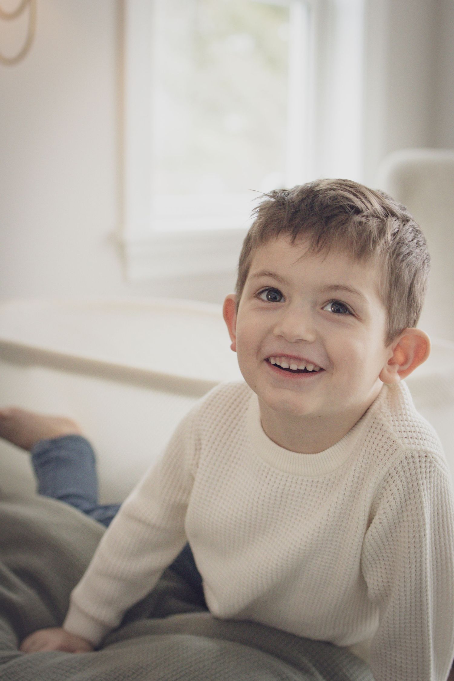 Andover family photographer capturing a joyful indoor portrait of a young child in a cozy white sweater smiling in soft natural window light
