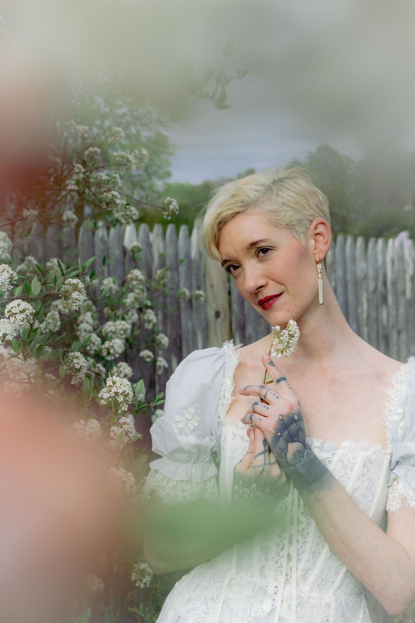 Close‑up portrait of a woman with short blonde hair wearing a vintage white lace dress, surrounded by blooming spring branches in a garden.