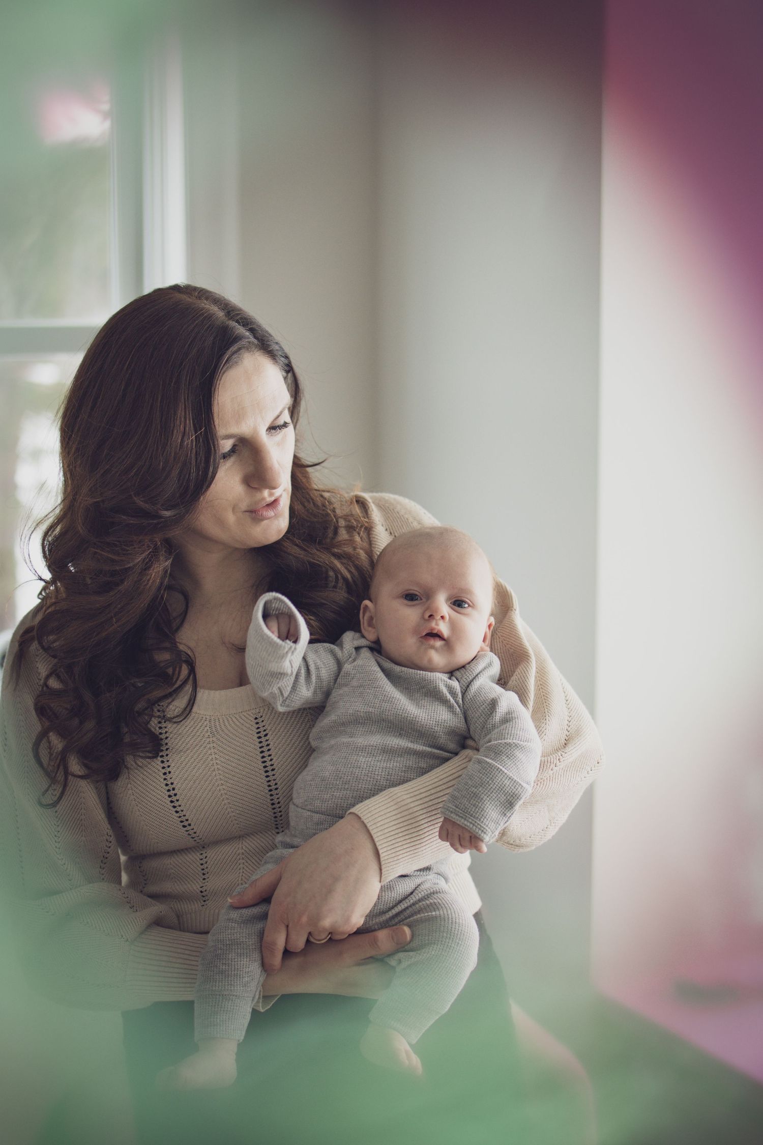 Mother holding her baby while looking into a mirror during a soft, natural indoor portrait session with warm, cozy light.