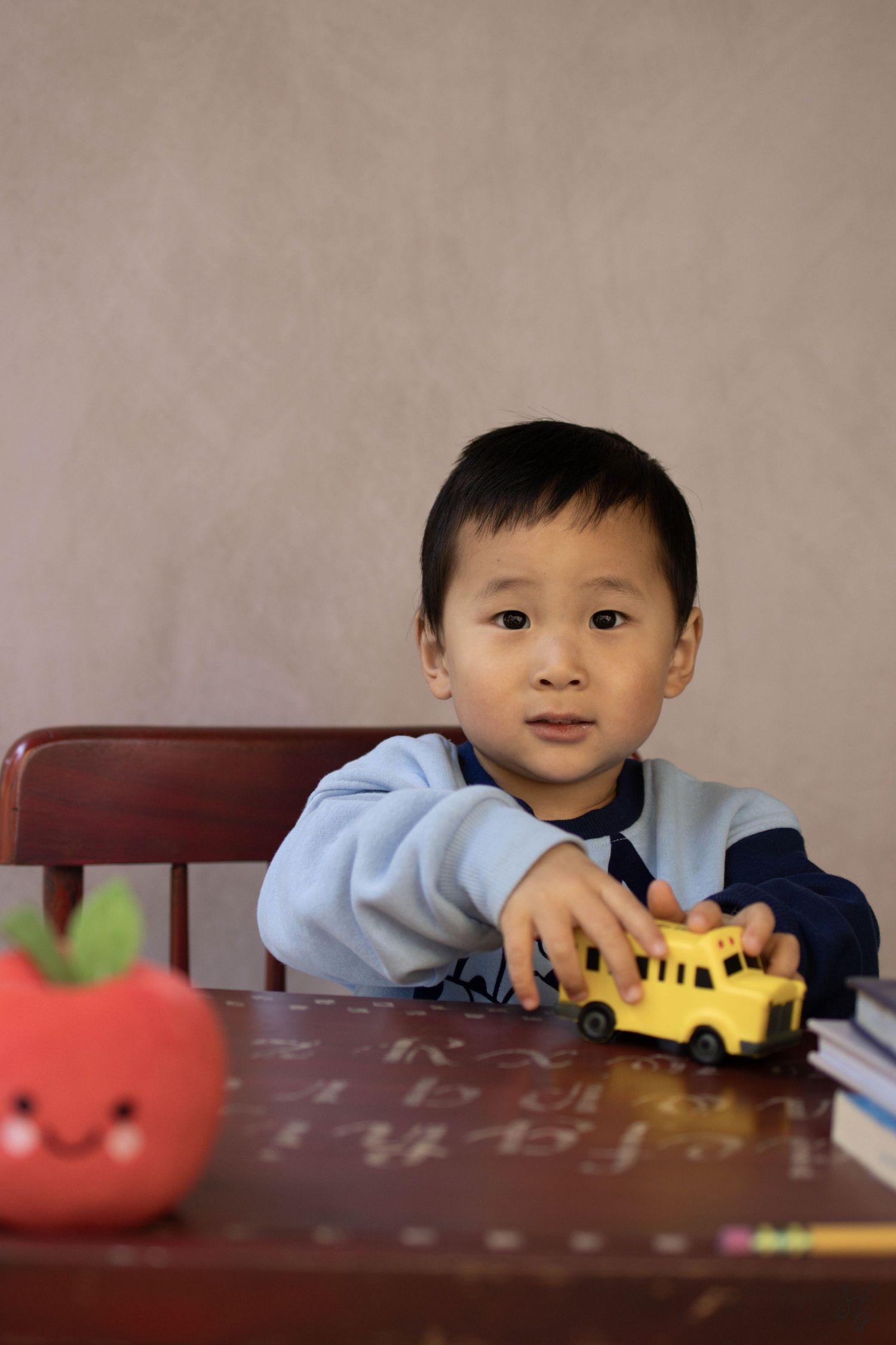 Young child sitting at a desk during a school portrait session, smiling while writing, with school‑themed props including books.