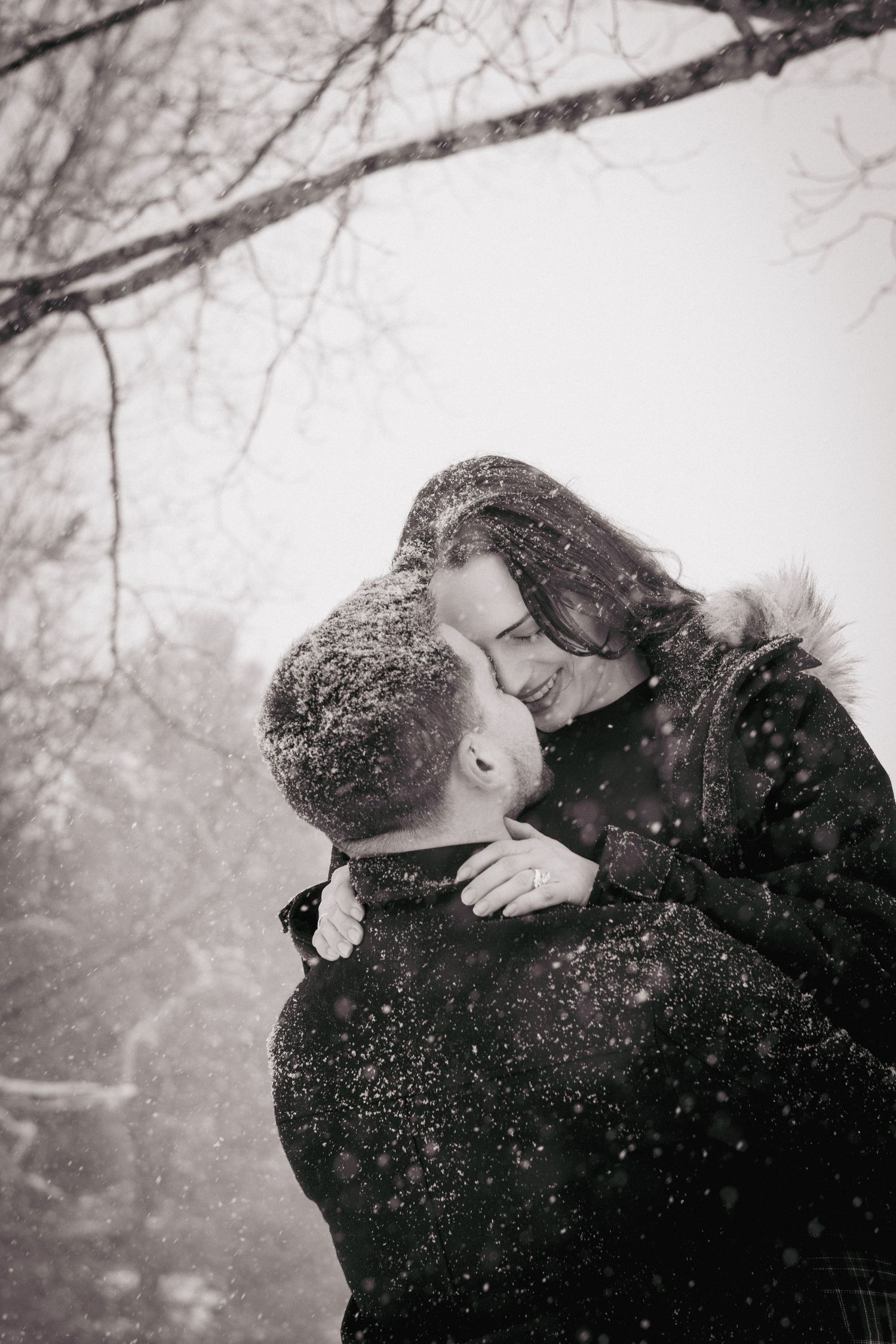 Couple embracing outdoors in falling snow, foreheads touching during a cozy winter portrait session.