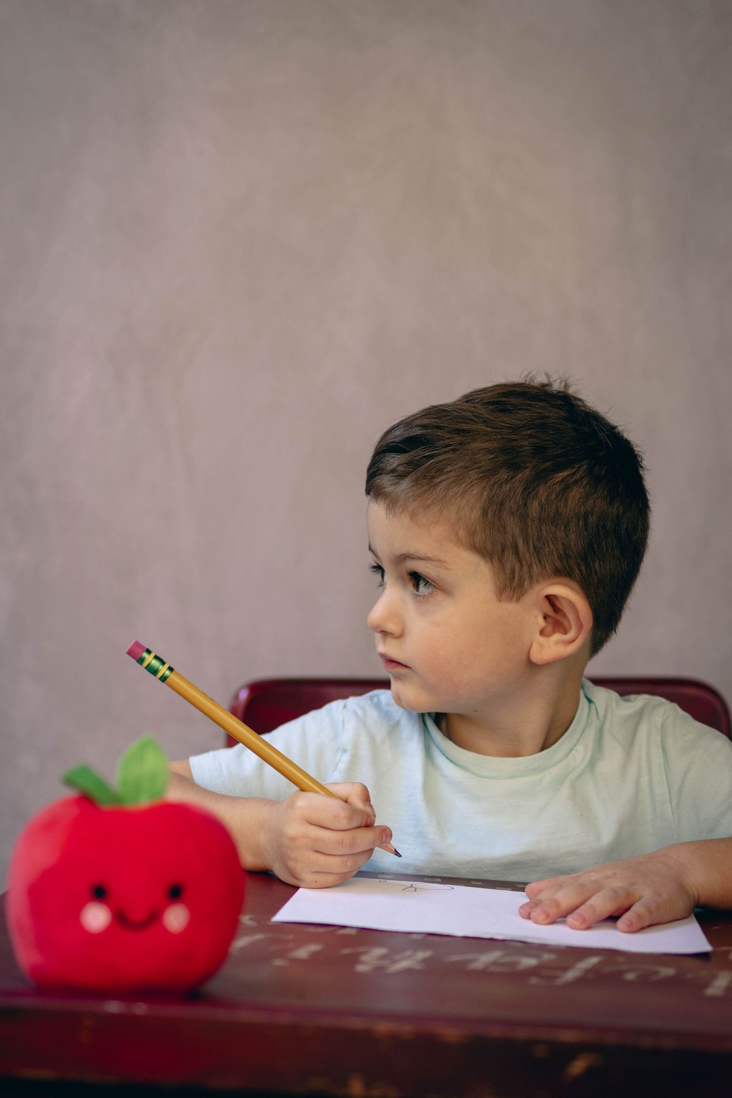 Young child sitting at a desk during a school portrait session, smiling while writing, with school‑themed props including books.