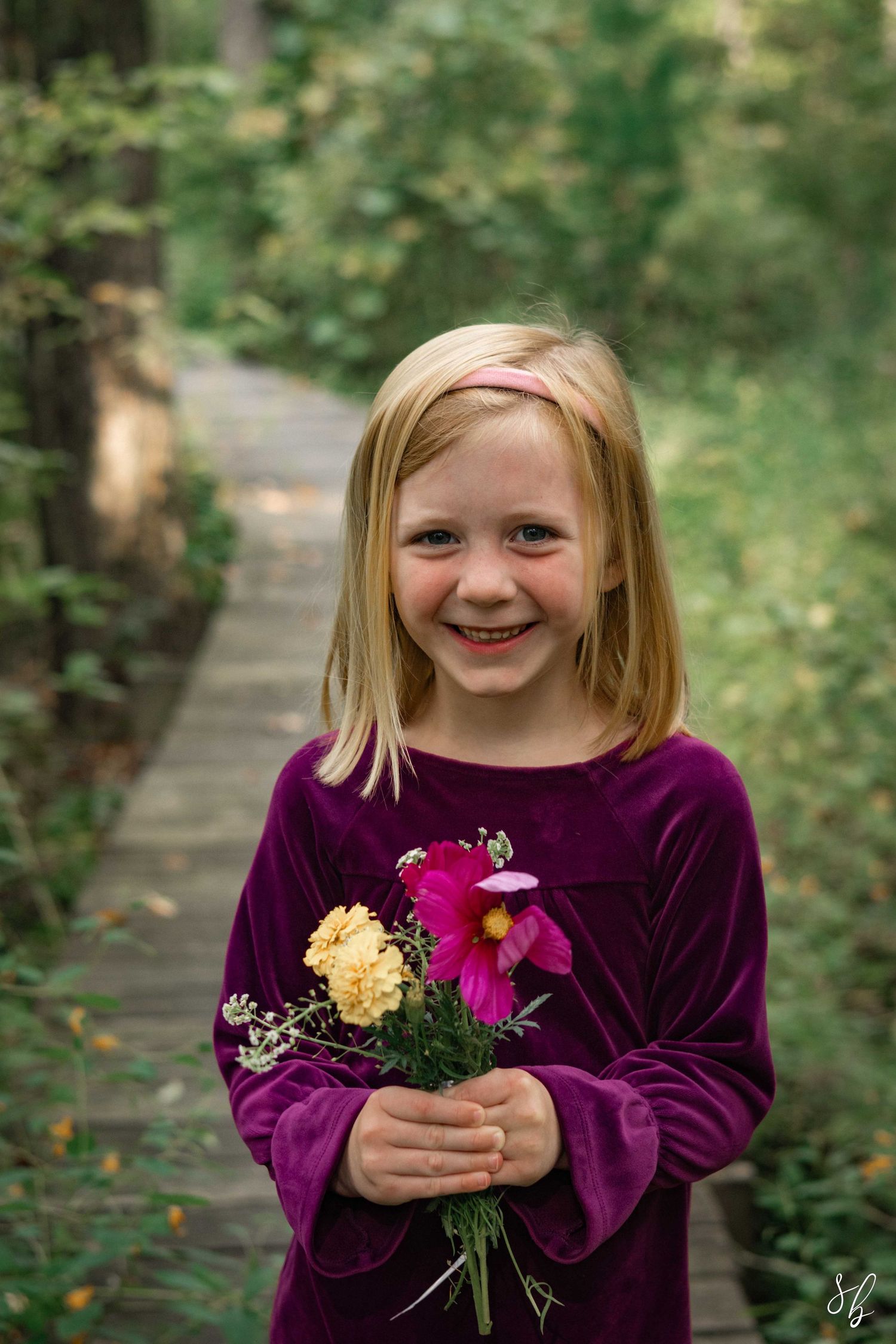 Andover family photographer capturing a young child in a purple velvet dress holding a bright flower bouquet on a wooden path in Andover.