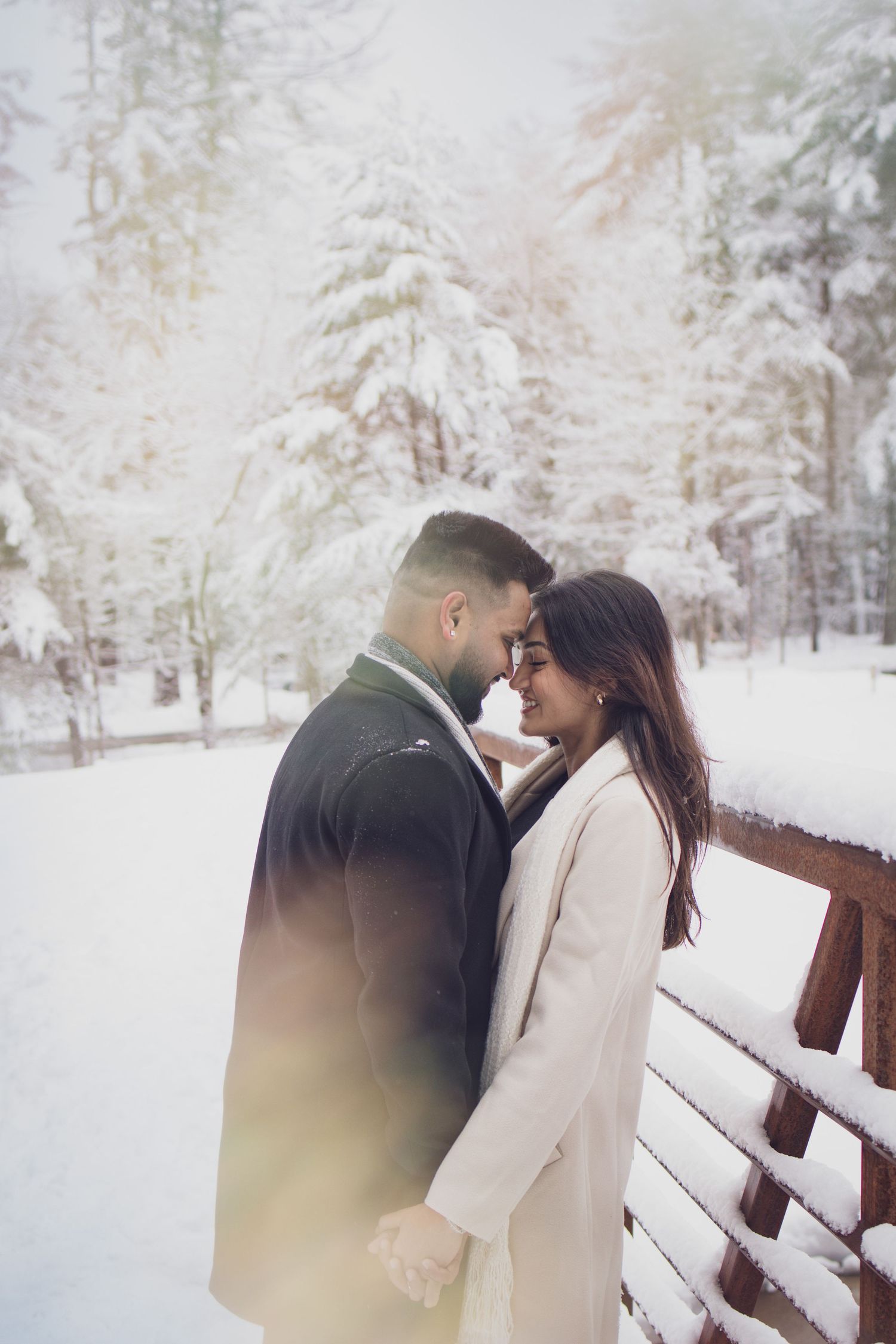 Couple embracing outdoors in falling snow, foreheads touching during a cozy winter portrait session.